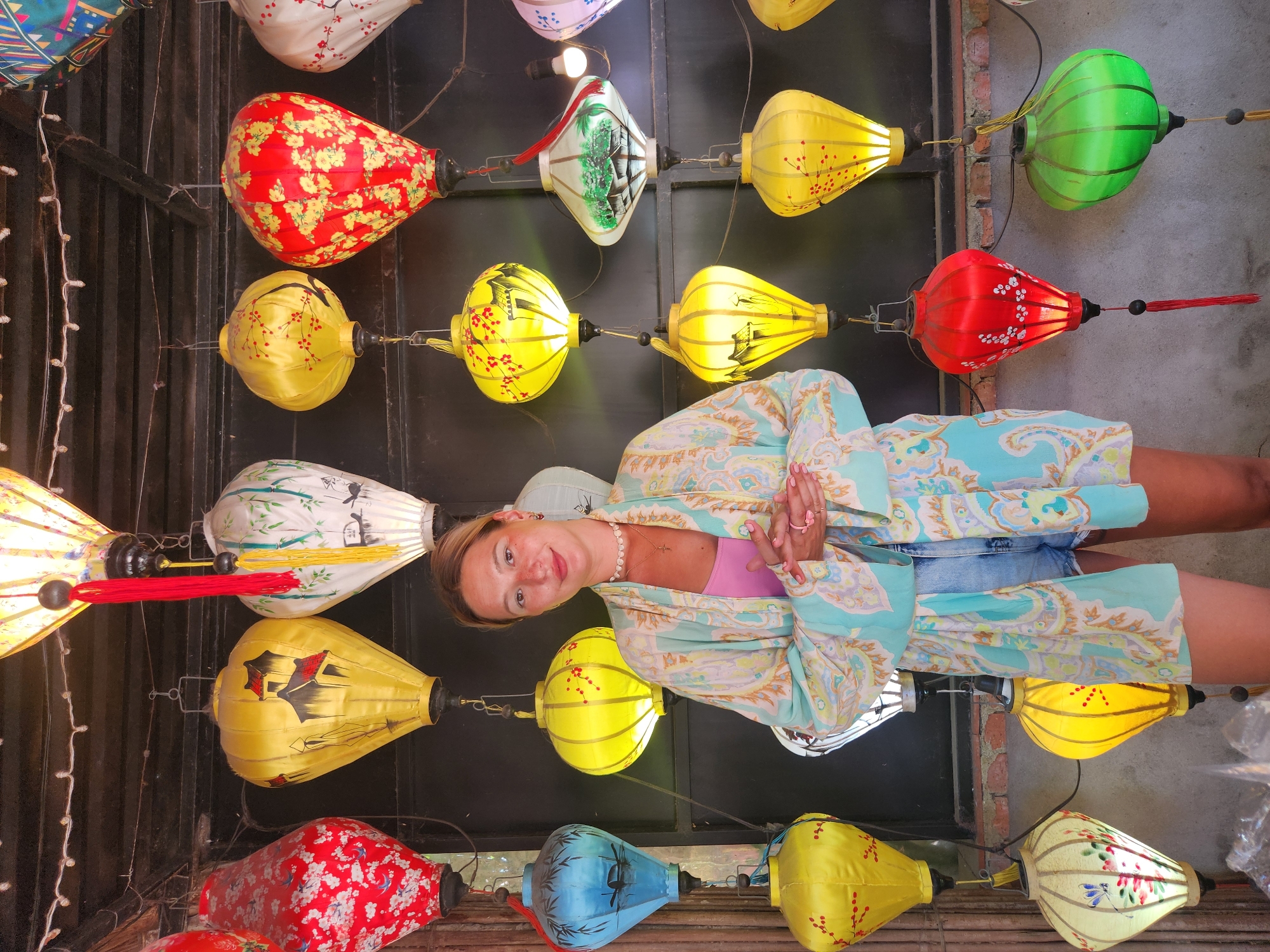 A woman stands in front of a wall adorned with numerous colorful, illuminated silk lanterns. She is wearing a light blue and beige patterned kimono-style robe over a pink tank top and denim shorts. Her hands are clasped in front of her. The lanterns are in various shapes and colors, including red, yellow, blue, green, and white, many featuring painted designs of landscapes and flora. The scene appears to be an outdoor market or shop in Hội An, Vietnam, a city known for its lantern-making tradition. The lanterns are hung at different heights and are illuminated from within, casting a warm glow.