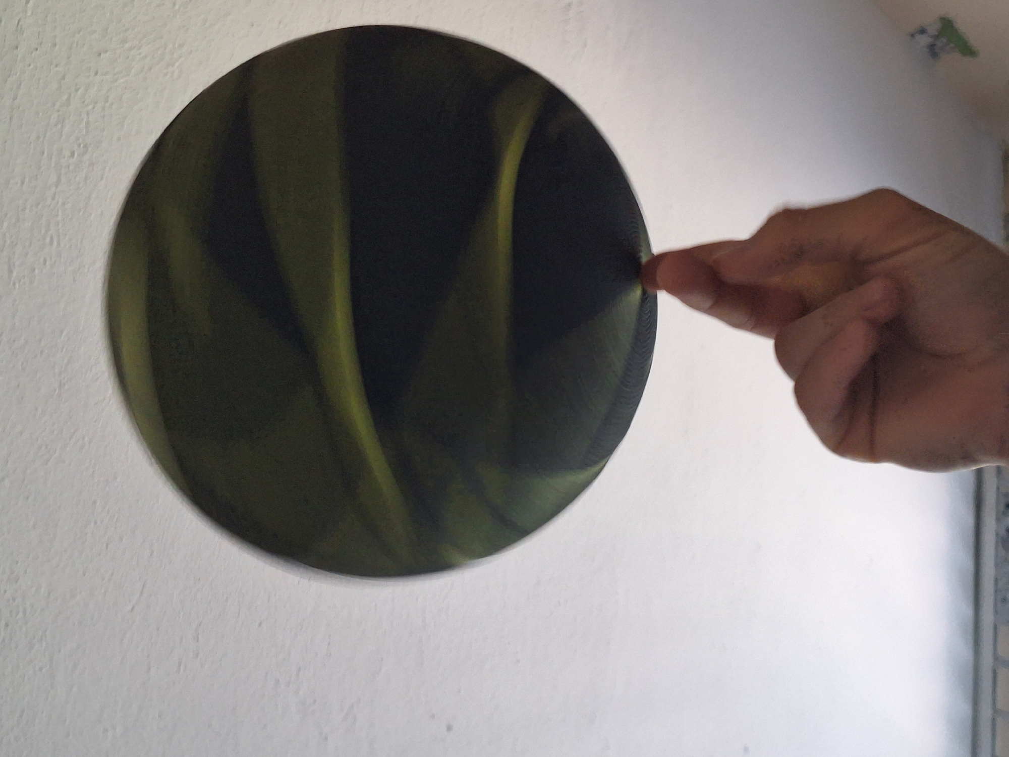 An adult's hand is balancing a dark green, circular object, possibly a frisbee or a large, flat plate, on the tip of their index finger. The object exhibits swirling patterns of lighter green and black. The background is a white, textured ceiling. The scene appears to be indoors, with no visible elements to indicate a specific city or country.