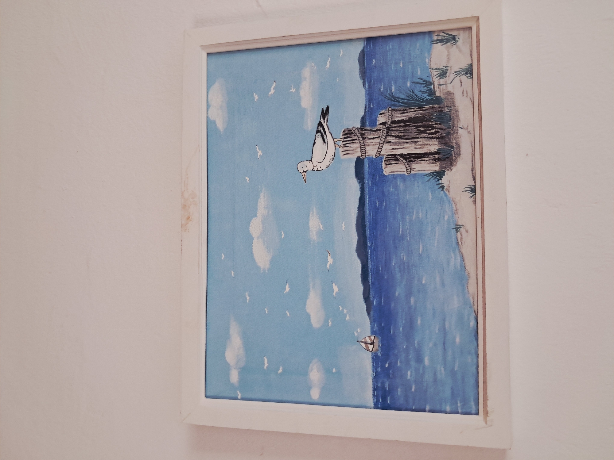A framed painting of a seascape hangs on a white wall. The painting depicts a pale blue sky with white clouds and seagulls. A body of water is rendered in various shades of blue with a single sailboat. A gray, wooden piling stands in the foreground with a white seagull perched on it. The top of the painting shows a sandy area with grasses. This is likely intended to depict a coastal scene near Juan Griego, Venezuela.