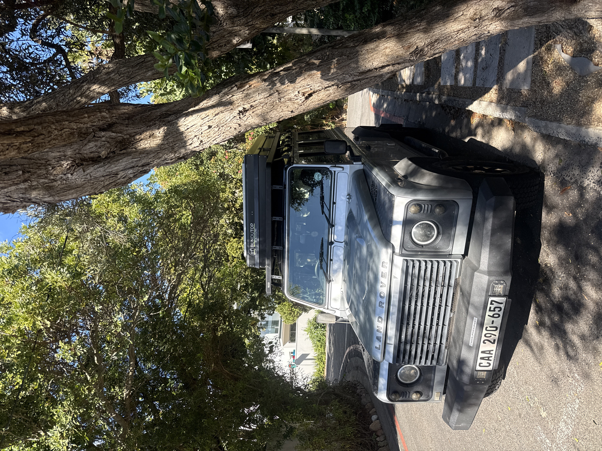 The image displays a grey Land Rover Defender 110, an off-road vehicle, parked outdoors under significant tree cover. The vehicle is positioned on the right side of the frame, facing slightly left, with its front grill prominently featuring the "LAND ROVER" brand. A black roof rack holds a "BUNDUTOP" tent system, indicating its adventure-ready setup. The front license plate clearly shows the South African registration "CAA 290-657", confirming the location context of Hout Bay, South Africa. A partial decal below the license plate reads "WILDPHOUNDS".
The setting is a treelined area, possibly a street or driveway, with large tree trunks and branches casting deep shadows across the vehicle and the ground. Patches of bright sunlight illuminate parts of the scene, suggesting a clear, sunny day, likely during late morning or early afternoon. Lush green foliage and dense tree canopies fill the background and upper left, with glimpses of a clear blue sky through the leaves. The ground comprises a paved surface, a concrete curb, and stepping stones leading away from the vehicle. A white building is partially visible through the trees in the distance. No people or specific activities are depicted; the scene conveys a sense of stillness and readiness for exploration.