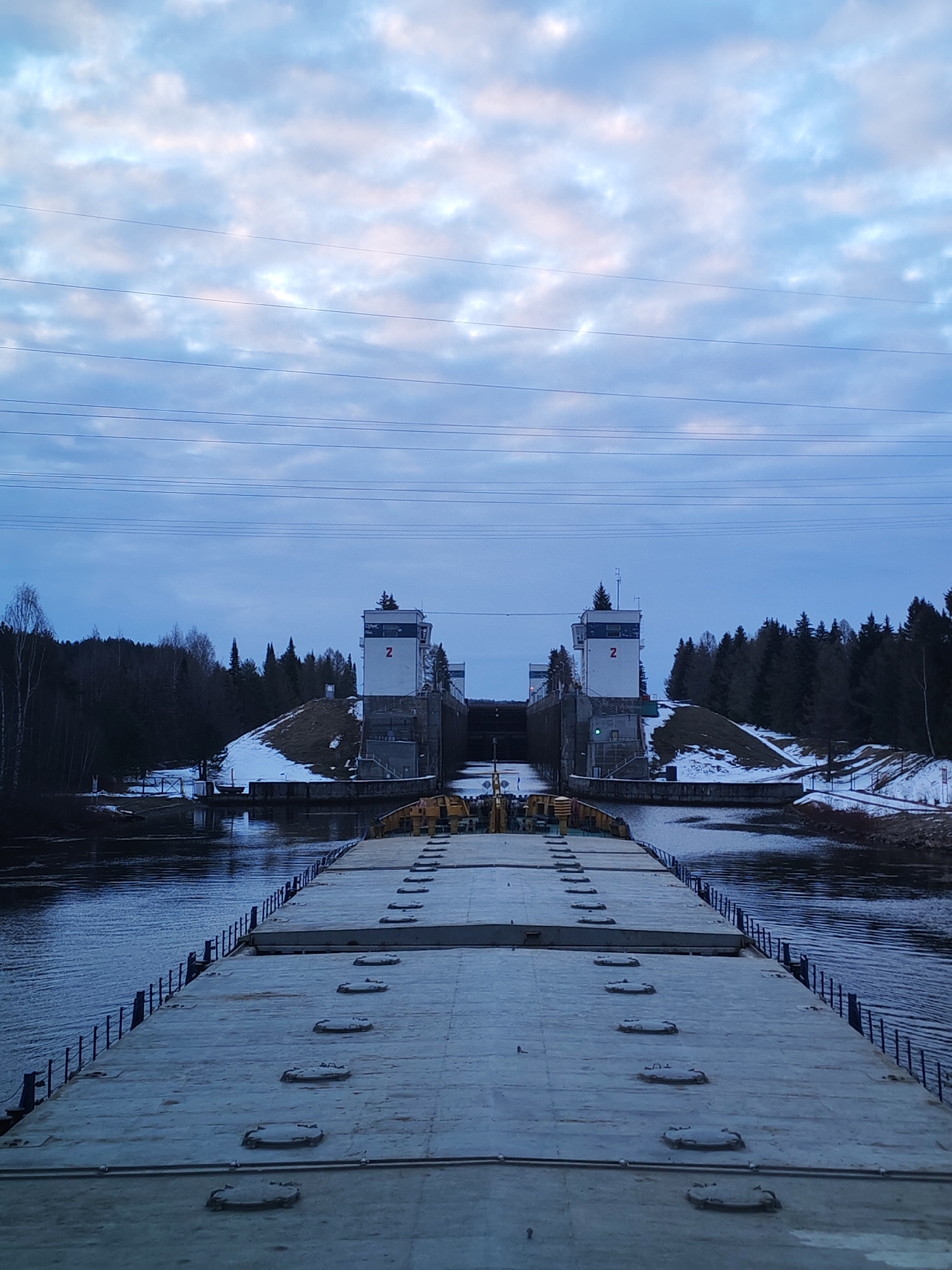The image presents a serene view of a canal lock system from the deck of a large vessel, likely a barge. In the foreground, the flat, grey deck of the vessel is visible, featuring multiple rows of oval and circular hatches or covers, some lightly dusted with snow. A low, dark railing runs along the vessel's port side, bordering the dark, rippling canal water. Yellow equipment is partially visible at the far end of the deck.
The main subject in the midground is a canal lock, distinguished by two prominent, light-colored control towers. Each tower displays a red number "2" on its side, signifying "Lock 2." These structures are flanked by low, snow-dusted hills adorned with dense evergreen forests, suggesting a cold climate, likely winter or early spring.
The background is dominated by an expansive, overcast sky, filled with layers of soft blue and white clouds, with subtle hints of pink, indicating either early morning or late afternoon light. Several parallel power lines stretch horizontally across the sky above the lock. The overall scene depicts a functional waterway, with the vessel either approaching or preparing to enter the lock, a common operation in canal navigation. The location is consistent with waterways found near Анхимово, Russia, such as sections of the Volga-Baltic Waterway. The atmosphere is quiet and functional, reflecting the cold, subdued light of the day.