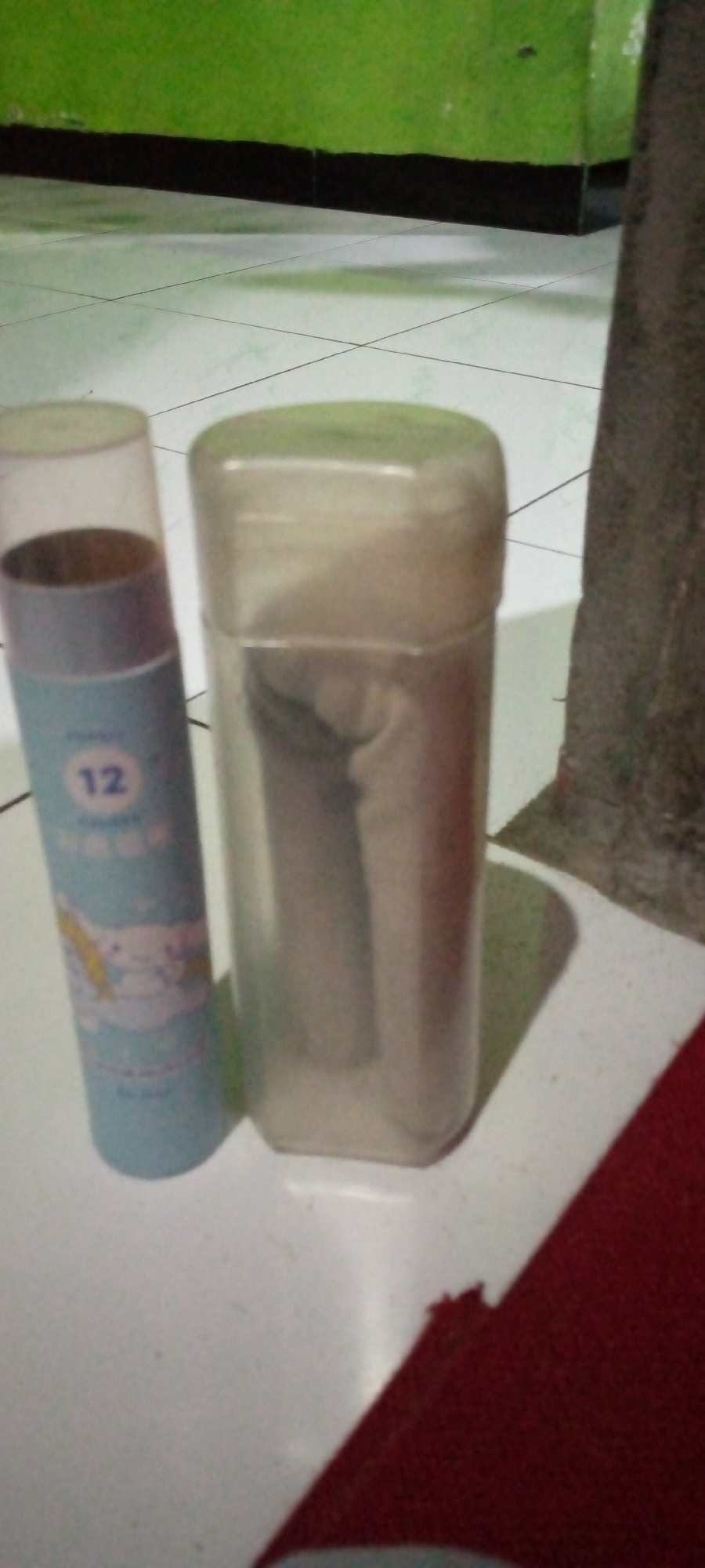 In the city of Sumberwono, Indonesia, two cylindrical containers are placed on a white tiled floor. The container on the left is light blue and has a partially visible transparent cap. It features a white graphic of a character, possibly a cartoon, and the number "12" in blue. The container on the right is a translucent plastic cylinder with a cap, appearing to contain a rolled-up object. The background consists of a green wall with a black border along the bottom. A dark, textured object, possibly a tree trunk or a pillar, is visible to the right. A red surface is partially visible in the foreground at the bottom right of the image.