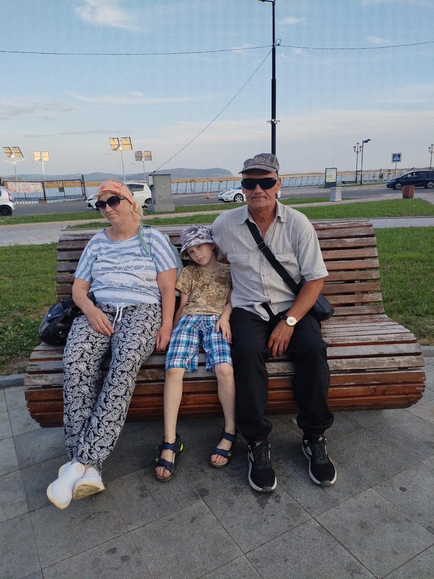 In Komsomolsk-on-Amur, Russia, three individuals are seated on a wooden slatted bench. The bench is positioned outdoors on a paved area adjacent to a grassy embankment. On the left, a woman wearing a light blue horizontally striped t-shirt and black patterned pants with white paisleys sits with her left leg extended forward. She has blonde hair tied back, wears dark sunglasses, and a peach-colored bandana on her head. Next to her, a young boy with light brown hair sits wearing a camouflage-patterned t-shirt and blue and white plaid shorts. He wears dark sandals. On the right, a man with short gray hair and a gray baseball cap sits with his legs crossed. He wears a light-colored short-sleeved button-down shirt, black pants, and black athletic shoes. He has dark sunglasses on and a black bag slung across his right shoulder. A watch with a gold face is visible on his left wrist. Behind the bench, a body of water, possibly a river, is visible, with a roadway running alongside it. Several cars are parked or driving on the roadway. Tall lampposts with multiple square light fixtures are spaced along the roadside. The sky above is a pale blue with scattered white clouds, and the overall lighting suggests it is late afternoon or early evening.