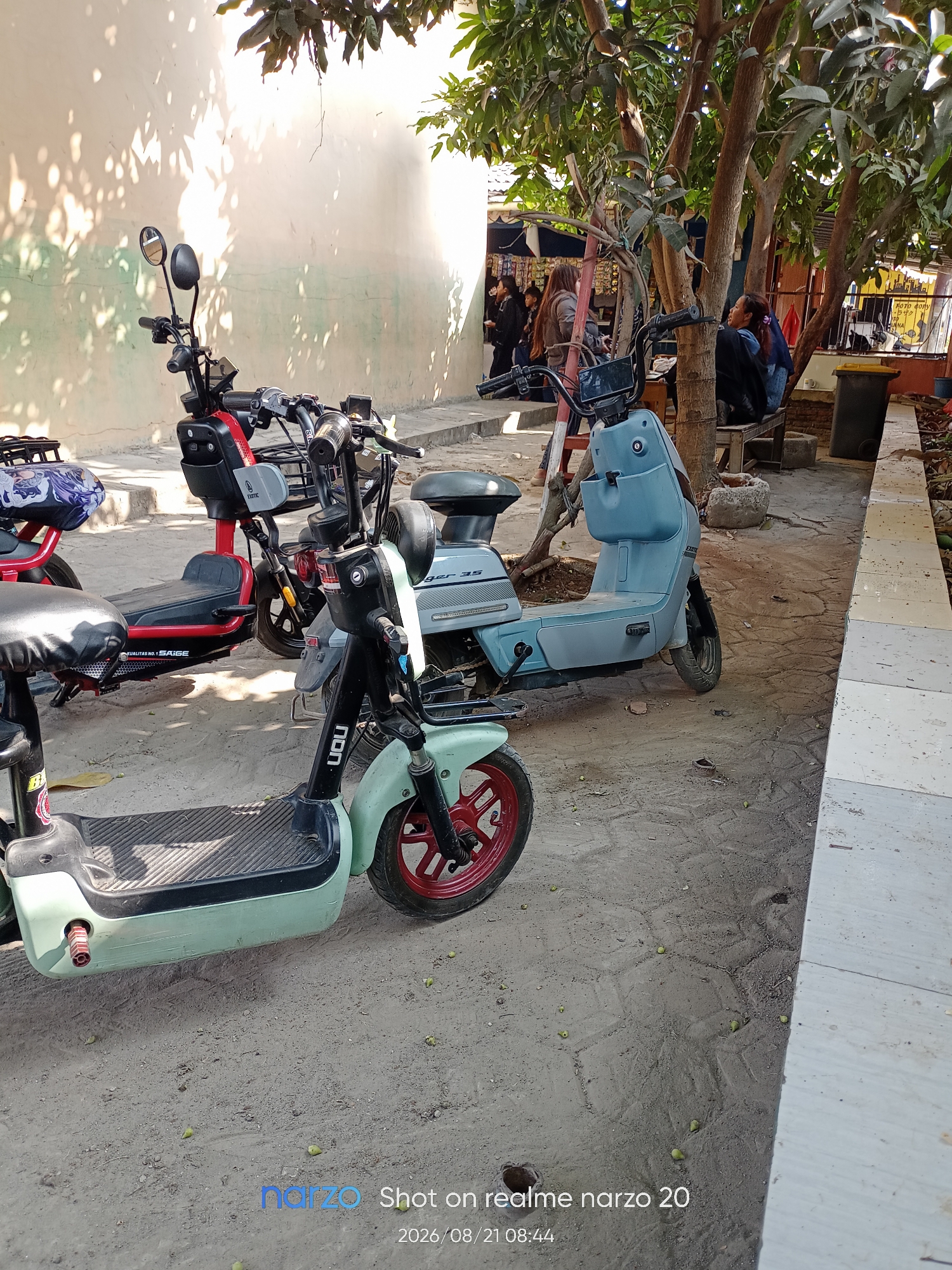 The image depicts a street scene in Karawang, Indonesia, likely a pedestrian walkway or sidewalk area. The foreground features two electric scooters parked side-by-side. The scooter on the left is mint green with a black seat and red wheel rims. The scooter on the right is light blue with a black seat and a basket. Behind the scooters, a light-colored building wall is visible, with dappled sunlight filtering through the leaves of trees overhead, casting shadows on the wall and ground. Further in the background, several individuals are seen under trees, some appearing to be seated or standing. There are also some structures that suggest a market or small shops. The ground is paved with textured concrete or pavers, and there is some debris on the ground, including small green fruits. The overall impression is a somewhat casual outdoor gathering or waiting area in an urban or semi-urban environment.