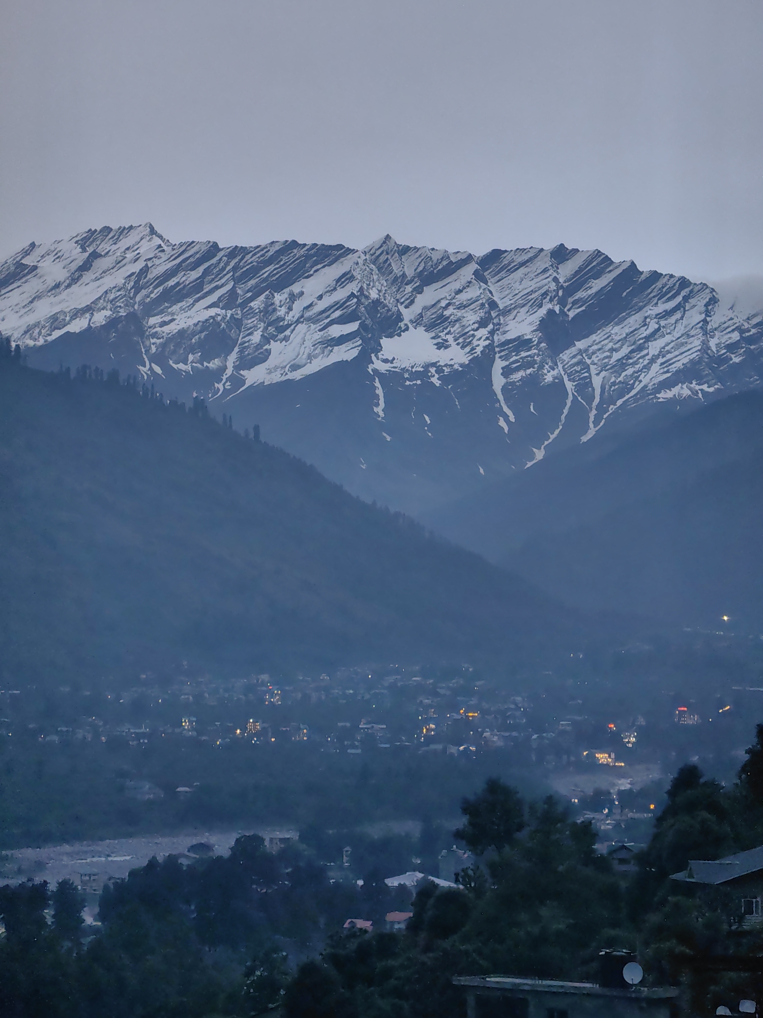 The image captures a panoramic view of a mountainous landscape at dusk, likely from a vantage point overlooking the town of Vashisht, India. The prominent feature is a range of snow-capped mountains that dominate the upper half of the frame. Their rugged peaks are sharply defined, with distinct patterns of snow contrasting against dark rock formations.
Below the mountains, a valley unfolds, veiled in a soft, atmospheric haze. Within the valley, a town is visible, characterized by a cluster of buildings illuminated by warm, twinkling lights, suggesting it is evening and the town is coming alive with artificial illumination. The lights are scattered and varied in intensity, hinting at a density of structures and activity.
The foreground is occupied by the dark silhouettes of trees, their branches creating a textured screen that partially obscures the view of the town and valley. These trees add depth to the image and frame the scene. The overall lighting is dim, consistent with twilight, with the sky a pale, muted gray. The environment appears to be a remote, mountainous region, likely a tourist destination given the visible infrastructure and lighting within the town. There are no discernible people or specific actions depicted in the scene, which focuses on the landscape and the human settlement within it.