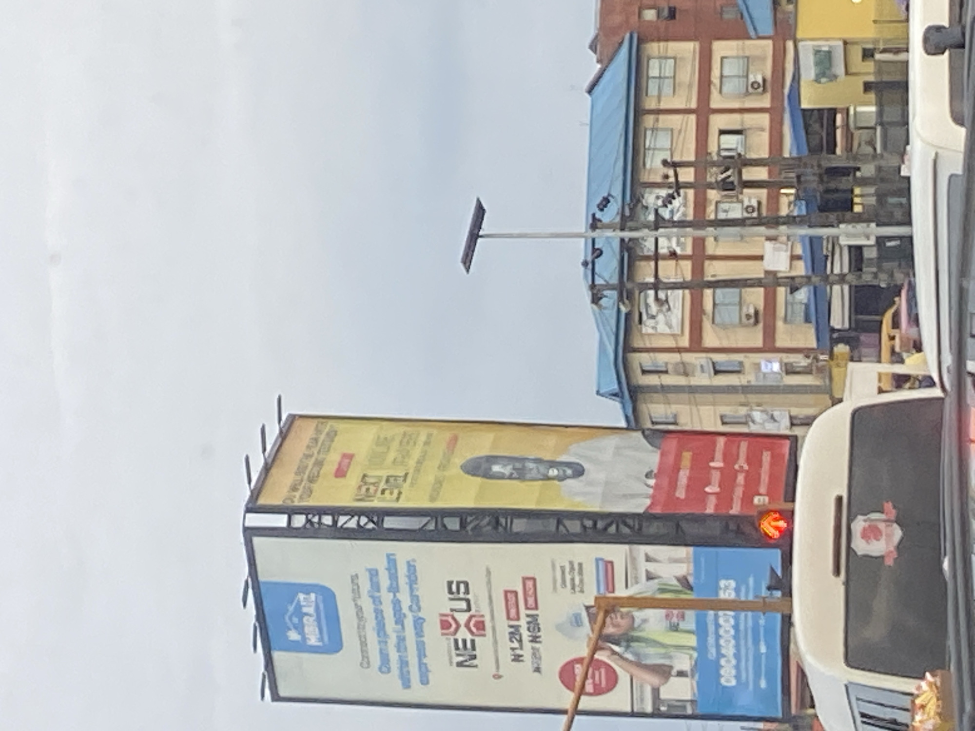 The image shows a street view in Ikorodu, Nigeria, with a prominent billboard dominating the scene. The billboard features advertisements for MBRAIZ and NEXUS. The left side of the billboard advertises "Connect to your future. Own a piece of land within the Lagos-Ibadan expressway Corridor." It also displays prices like "N1.2M" and "N2M". A worker wearing a hard hat and high-visibility vest is depicted on this section of the billboard. The right side of the billboard is yellow and black and features the text "NEXT LEVEL FAITHERS". A portrait of a man with dark skin, wearing a white shirt, is prominently displayed on this side.
Below the billboard, a traffic light shows a red arrow, indicating a stop signal. A white commercial vehicle, possibly a mini-bus or van, is in the foreground, partially obscuring the lower portion of the billboard and traffic light. The vehicle has a red and white emblem on its rear window. In the background, there are multi-story buildings with blue and yellow facades, and telephone poles with numerous wires. The sky is overcast. The overall impression is a busy urban environment with commercial advertising and traffic infrastructure.