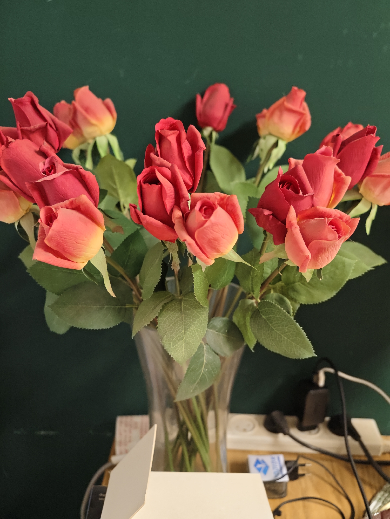 A bouquet of red and pink roses is arranged in a clear glass vase, with lush green leaves. The vase sits on a wooden surface in front of a dark green wall. To the right of the vase, a white power strip is plugged into an electrical outlet, with several black and white plugs connected. There are also some charging adapters and cables visible on the wooden surface. The lighting in the image is dim, creating a moody atmosphere. The scene appears to be indoors, likely in a home or office setting. No specific location in Petah Tikva, Israel, can be determined from the image.