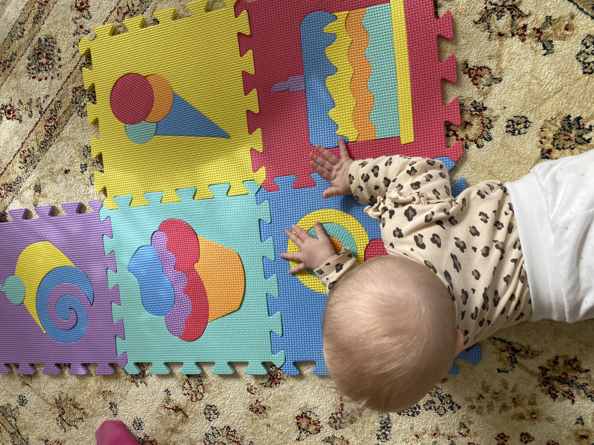 An overhead view captures a baby lying on their stomach on a colorful foam play mat, which covers a traditional patterned rug. The baby's head is visible from the back, showing light-colored hair. They are wearing a light-toned, long-sleeved top with a leopard print pattern, and a white garment is visible on their lower body. The baby's small hands are resting on the play mat, suggesting interaction with the surface.
The foam play mat is made up of several interlocking tiles in bright colors such as yellow, pink, red, blue, light blue, and purple. Each tile features different cutout shapes. Notable designs include an ice cream cone with scoops on a yellow tile, wavy patterns in various colors on a red tile, and spiral shapes with circles on a purple tile. Other tiles show abstract forms and concentric circles. The foam surface has a textured, grid-like pattern.
The play mat is situated on a cream or beige rug adorned with intricate floral and geometric designs in shades of red, blue, and brown, visible where the mat does not cover it. A small pink object, possibly a baby's foot or sock, can be seen at the bottom left of the image. The scene depicts an indoor setting, likely a home, providing a stimulating and soft play area for the baby. There is no visible text.
