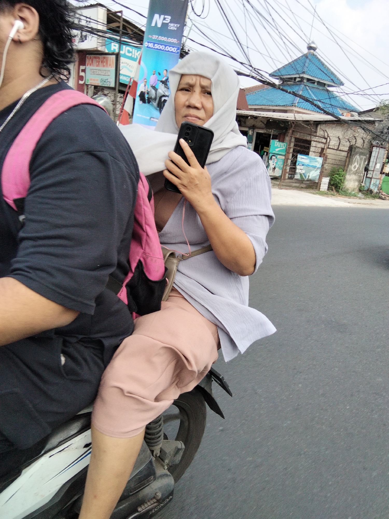 The image depicts two individuals on a motorcycle, likely in the city of South Tangerang, Indonesia, given the context. The person in the foreground, on the left, is wearing a dark shirt and a bright pink backpack. They have dark hair and are wearing white earbuds. The person behind them, on the right, is wearing a light grey tunic, light pink pants, and a light grey headscarf. They are holding a black smartphone in their right hand, with their left hand resting on their lap. A light brown strap, possibly from a bag, is visible across their chest. The background shows a street scene with buildings, including a structure with a blue tiled roof resembling a mosque, and a variety of overhead electrical wires. A banner with text and images is also visible. The road surface is asphalt.