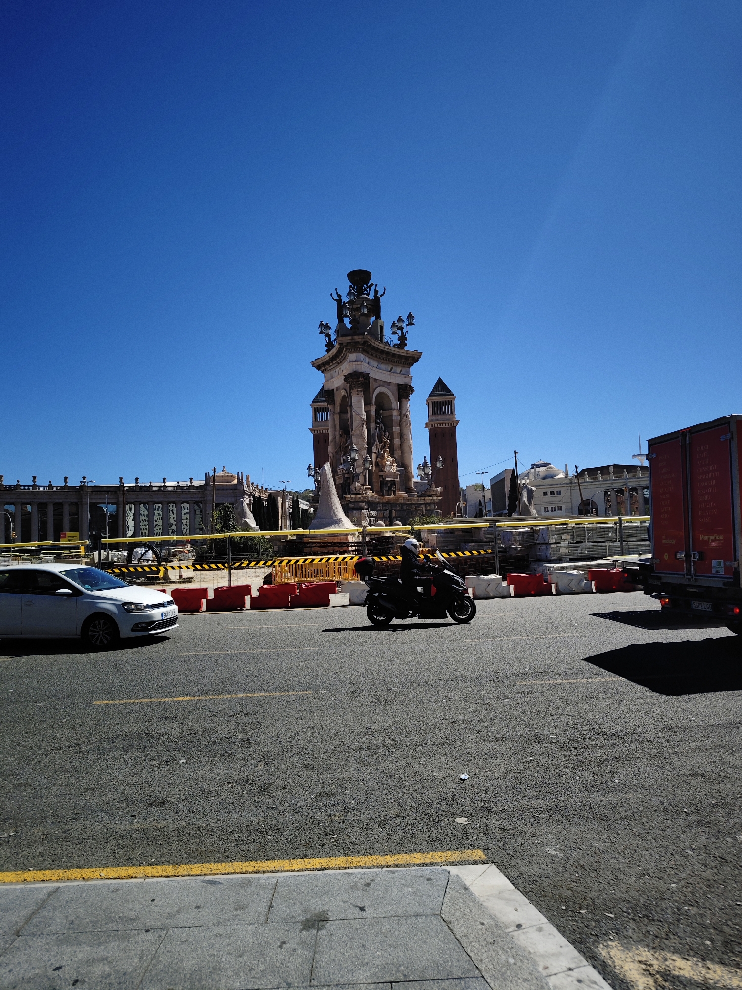 The image depicts the Plaça d'Espanya in Barcelona, Spain. The large fountain, Font de la Plaça d'Espanya, is visible in the background, surrounded by construction barriers. A white car is on the left side of the road and a black motorbike in the middle. A red truck on the right partially obscures the view. The sky is clear and blue.
