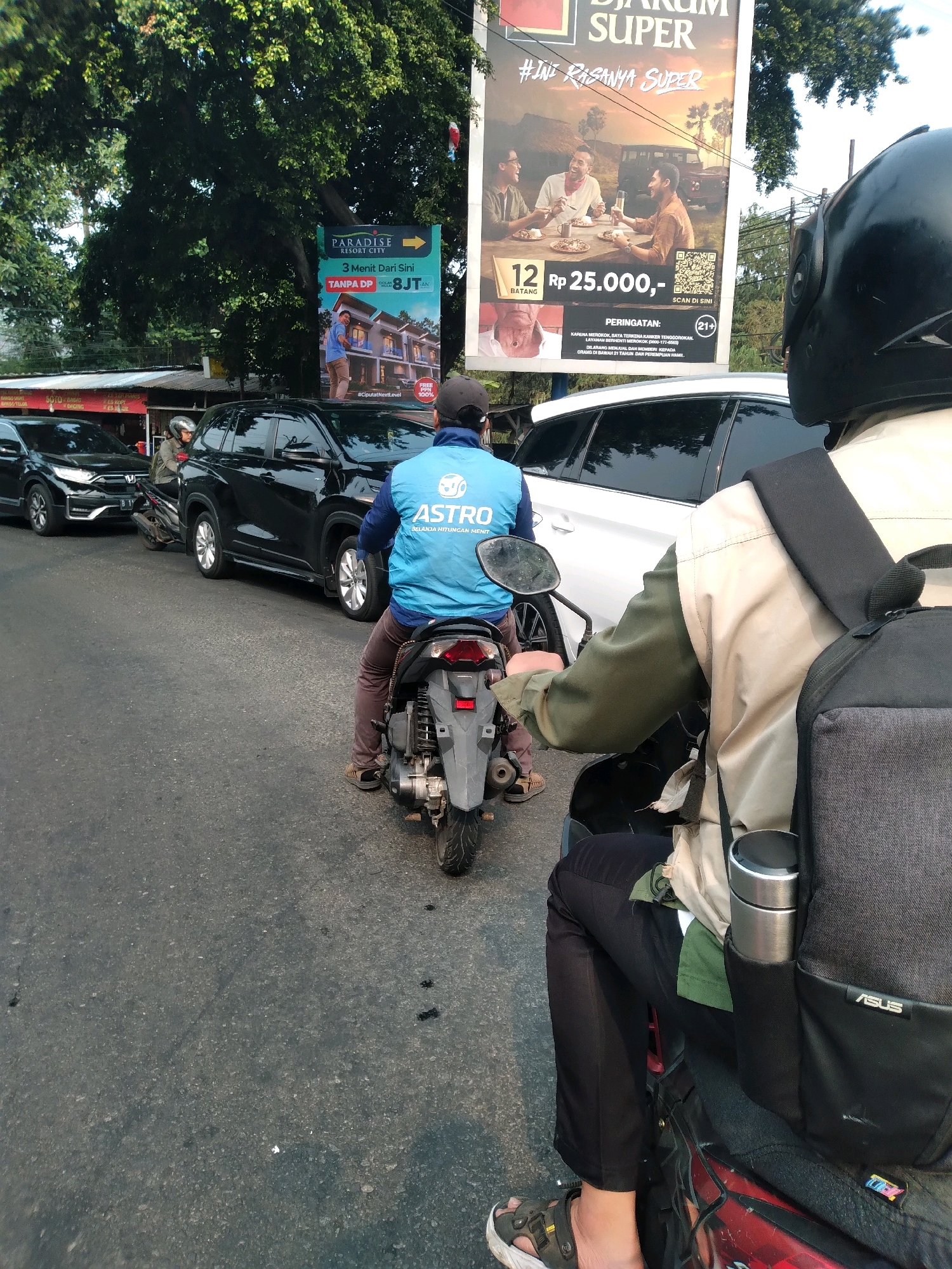 The image captures a street scene in South Tangerang, Indonesia, likely during daylight hours. In the foreground, the back of a motorcyclist wearing a blue vest with the "ASTRO" logo and a black helmet is visible, indicating they are operating a motorcycle. To the right, another person on a motorcycle is partially visible, also wearing a helmet and a tan jacket over a green shirt, with a backpack containing a silver water bottle.
The street is lined with several vehicles, including black cars and SUVs, suggesting moderate traffic. In the background, a large billboard advertises "Djarum Super" with an image of people dining and text in Indonesian. Another billboard promoting "PARADISE RESORT CITY" is also visible, featuring an image of a residential property. Tall trees with lush green foliage border the street, suggesting a somewhat green urban environment. The road surface appears to be asphalt, with some dark marks possibly indicating oil or other debris. The overall impression is a typical urban street in Indonesia with billboards, traffic, and roadside vegetation.
