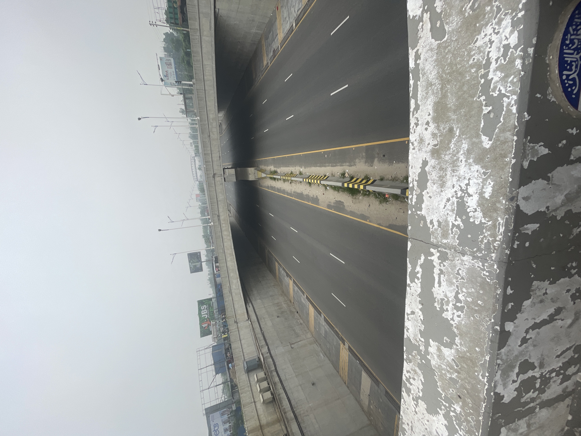 The image shows a multi-lane highway in Purbachal, Bangladesh. The highway is mostly empty, with white lane markings and yellow edge lines. A median with black and yellow striped barriers runs down the center. The roadway is under an overpass or flyover. The sky is overcast and hazy. A concrete barrier with peeling paint is in the foreground. A billboard with "JBS" is visible in the distance.