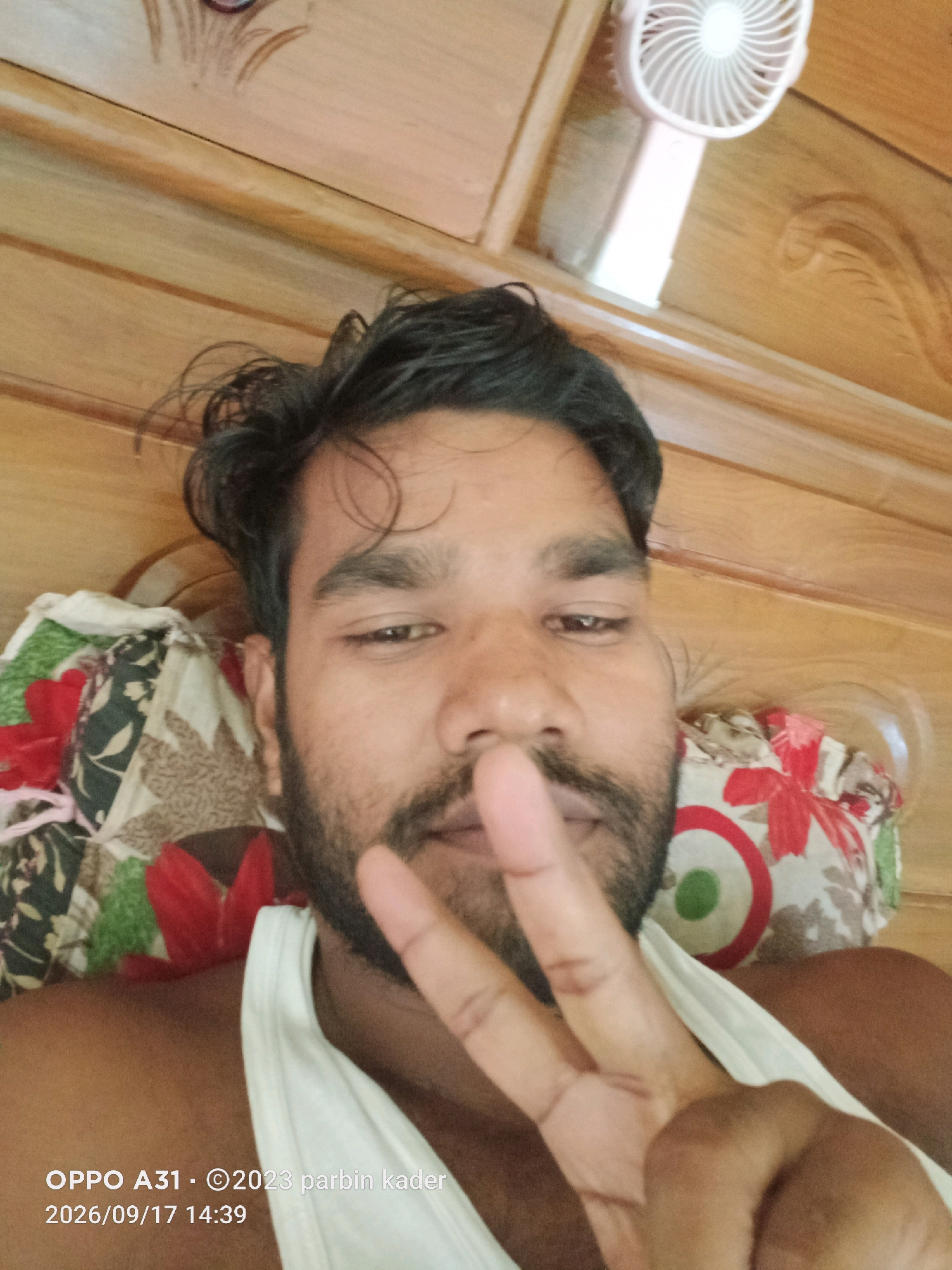 The image is a close-up, eye-level, selfie-style photograph of a man lying down. He has dark, messy hair, a beard, and is wearing a white tank top. He is holding up two fingers in a peace sign gesture towards the camera. The background consists of a wooden surface with decorative carvings and a patterned pillow. A small, white, portable fan is visible in the upper right corner of the image. The bottom of the image contains text overlay indicating "OPPO A31", a copyright notice for "2023 parbin kader", and a date and time "2026/09/17 14:39". The overall impression is that this is a casual personal photograph taken indoors. There is no discernible information in the image to suggest the location is in Chamuakhat, India.