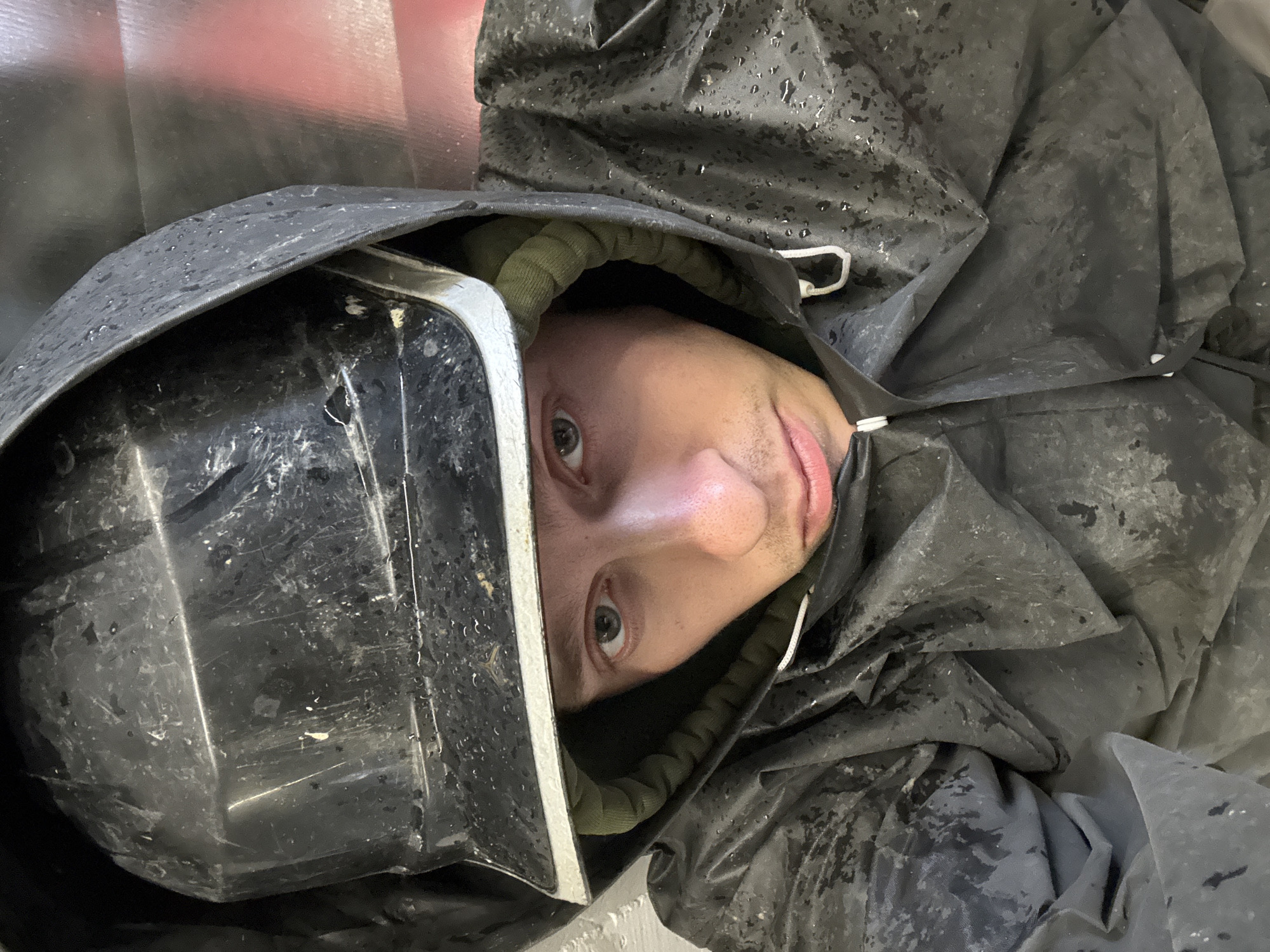 A close-up, high-angle selfie shows a young adult male wearing a black, hooded rain suit and a black helmet with a white trim. The subject's face is partially obscured by the helmet and hood. The rain suit appears to be wet, with visible water droplets on the fabric. The background is out of focus, but hints of red and gray suggest an industrial or urban environment. No specific Moscow landmarks are visible. The image is undated and unlocated.