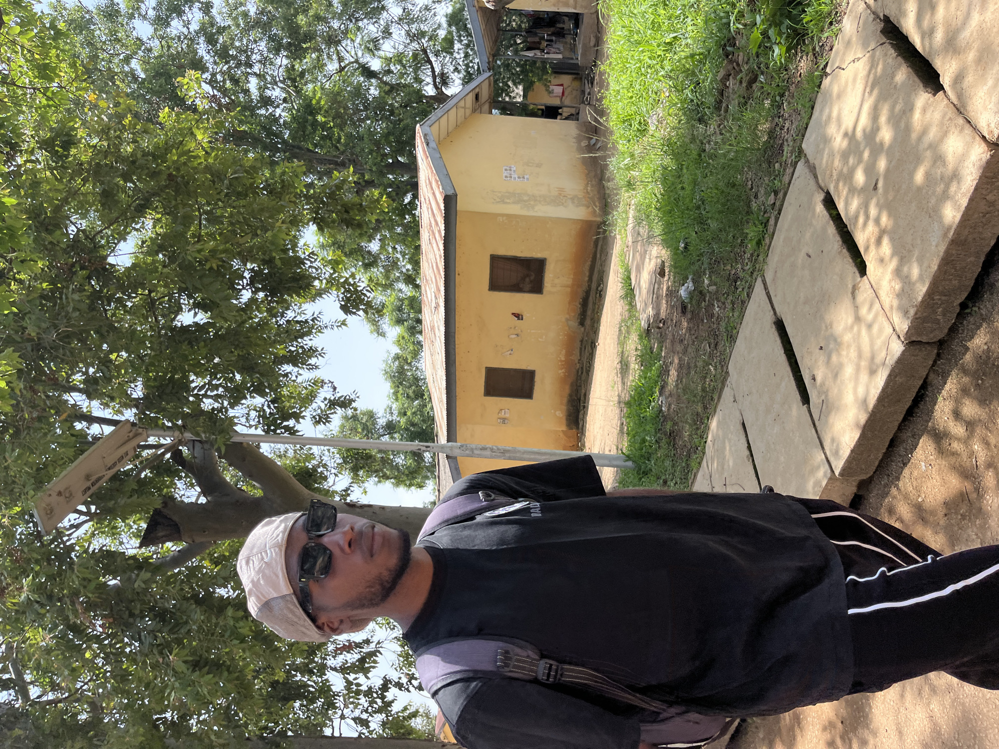 In Rafin Par, Nigeria, a young man is seen walking outdoors, wearing a black t-shirt, black sweatpants with white stripes, a light-colored baseball cap, and sunglasses. He carries a purple backpack. The background features a yellow, single-story building with a rusty metal roof and two dark-paned windows. Adjacent to this structure is another building with a covered outdoor area. Trees with green foliage and a bright blue sky are visible above. The ground is a mix of dirt, cracked concrete slabs, and lush green grass. A solar-powered street light stands near the building.