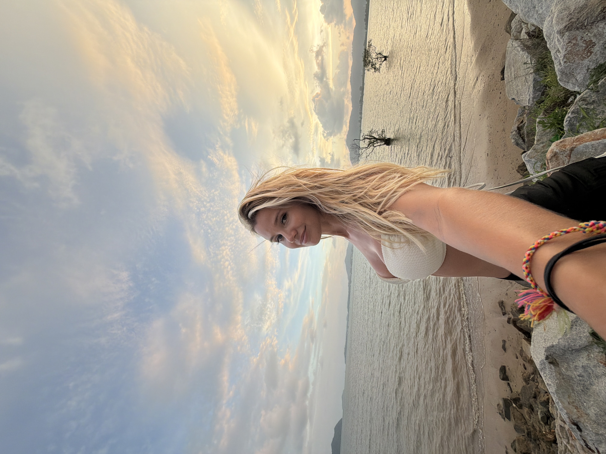 A woman with long, blonde hair, wearing a white strapless bikini top and black bottoms, is taking a selfie on a rocky shoreline with the ocean and a cloudy sky in the background. The image appears to have been taken at dusk, as indicated by the soft, warm light illuminating the sky. In the distance, two silhouetted trees stand in the water, and a mountain range is visible on the horizon. The woman is looking at the camera and smiling. The scene suggests a tranquil and picturesque coastal location, possibly in Wichit, Thailand, given the presence of tropical vegetation and the general aesthetic of the landscape.