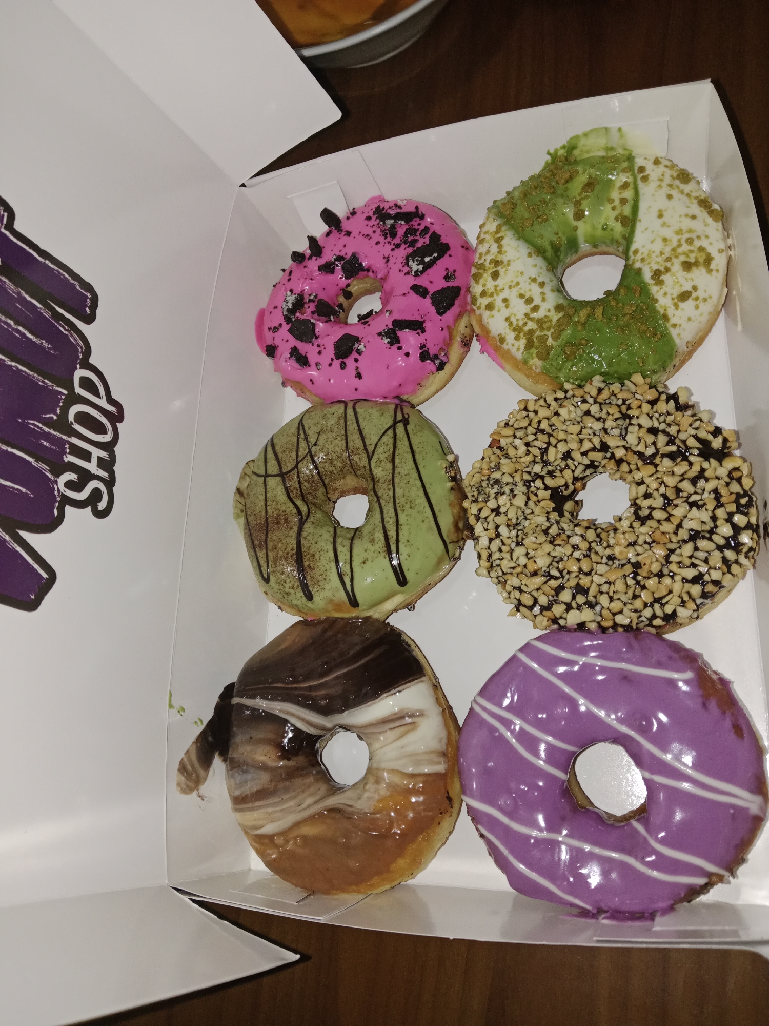 A box of six donuts sits on a wooden surface in Unknown, Unknown. The donuts are arranged in two rows of three. The top row consists of a pink donut with black sprinkles, a white donut with green frosting and crumbs, and the middle row has a light green donut with dark green drizzle and a donut covered in chopped nuts. The bottom row has a donut with chocolate and white icing, and a purple donut with white stripes. The box is white and has the words "SHOP" in a purple font on the side.