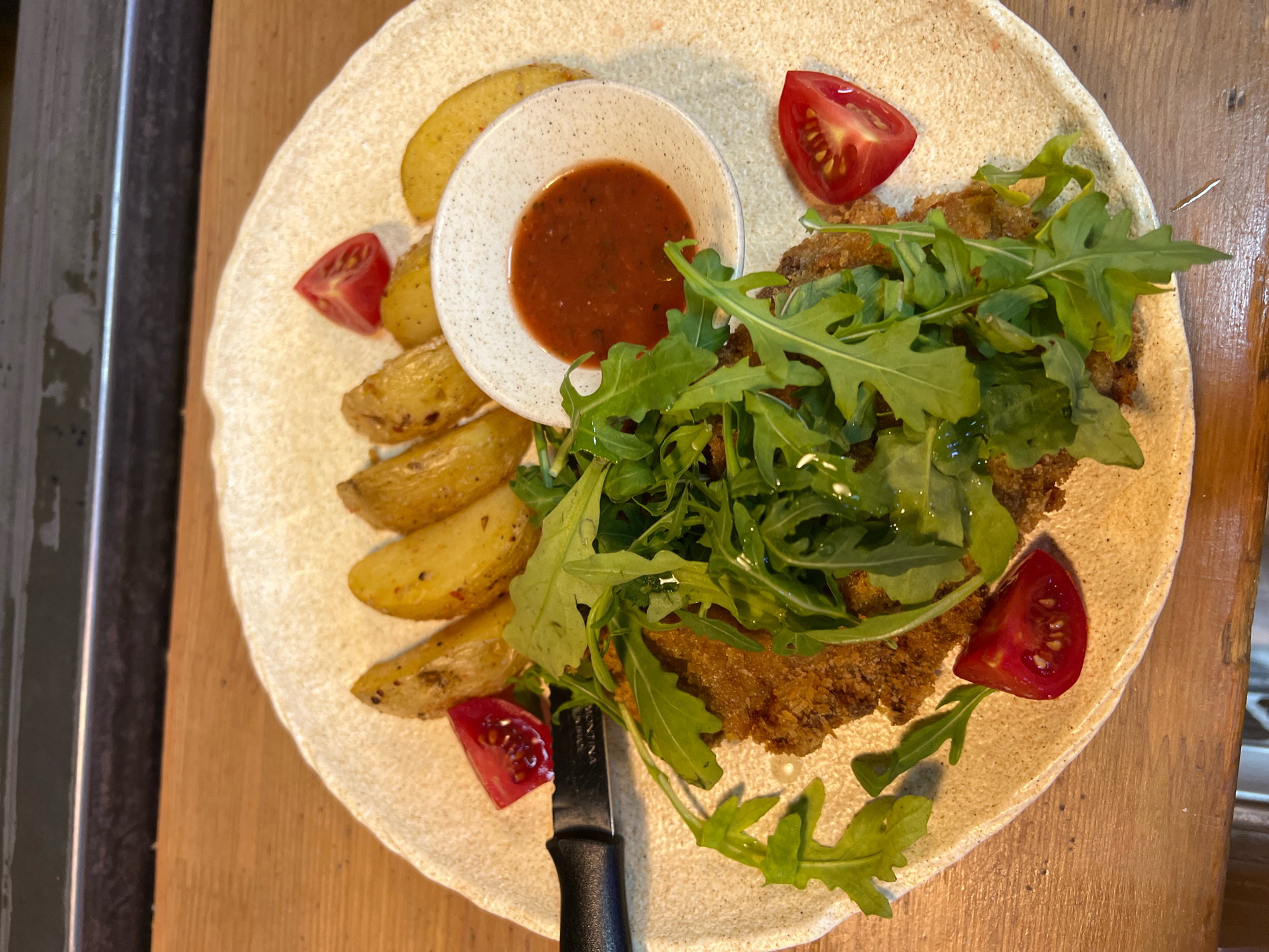 A close-up, overhead shot shows a plate of food on a wooden surface. The plate is round and speckled with a light brown and white pattern. It contains a breaded cutlet, a pile of rocket salad, several potato wedges, a small bowl of red sauce, and three wedges of cherry tomato. A knife with a black handle is positioned near the bottom of the plate. The overall lighting suggests an indoor setting, possibly a restaurant. There are no people visible in the image. The food appears to be ready to be eaten. The image is taken in Bishkek City, Kyrgyzstan.