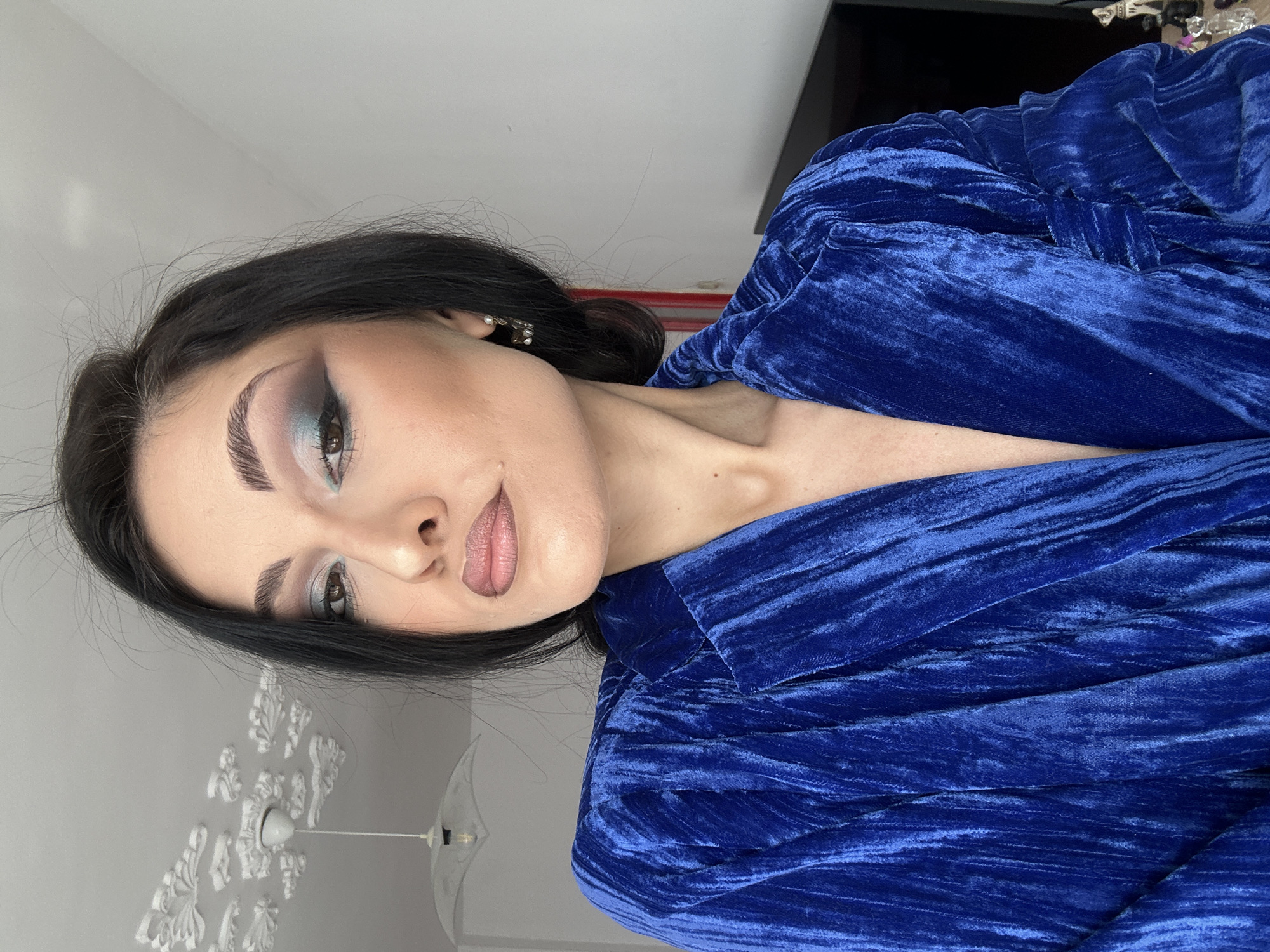 A portrait of a woman with dark hair, wearing a blue velvet garment, is positioned against a light-colored wall. She is partially facing the camera, her gaze directed towards the lower left corner of the frame. The background includes a decorative light fixture on the wall. The exact location is unknown.