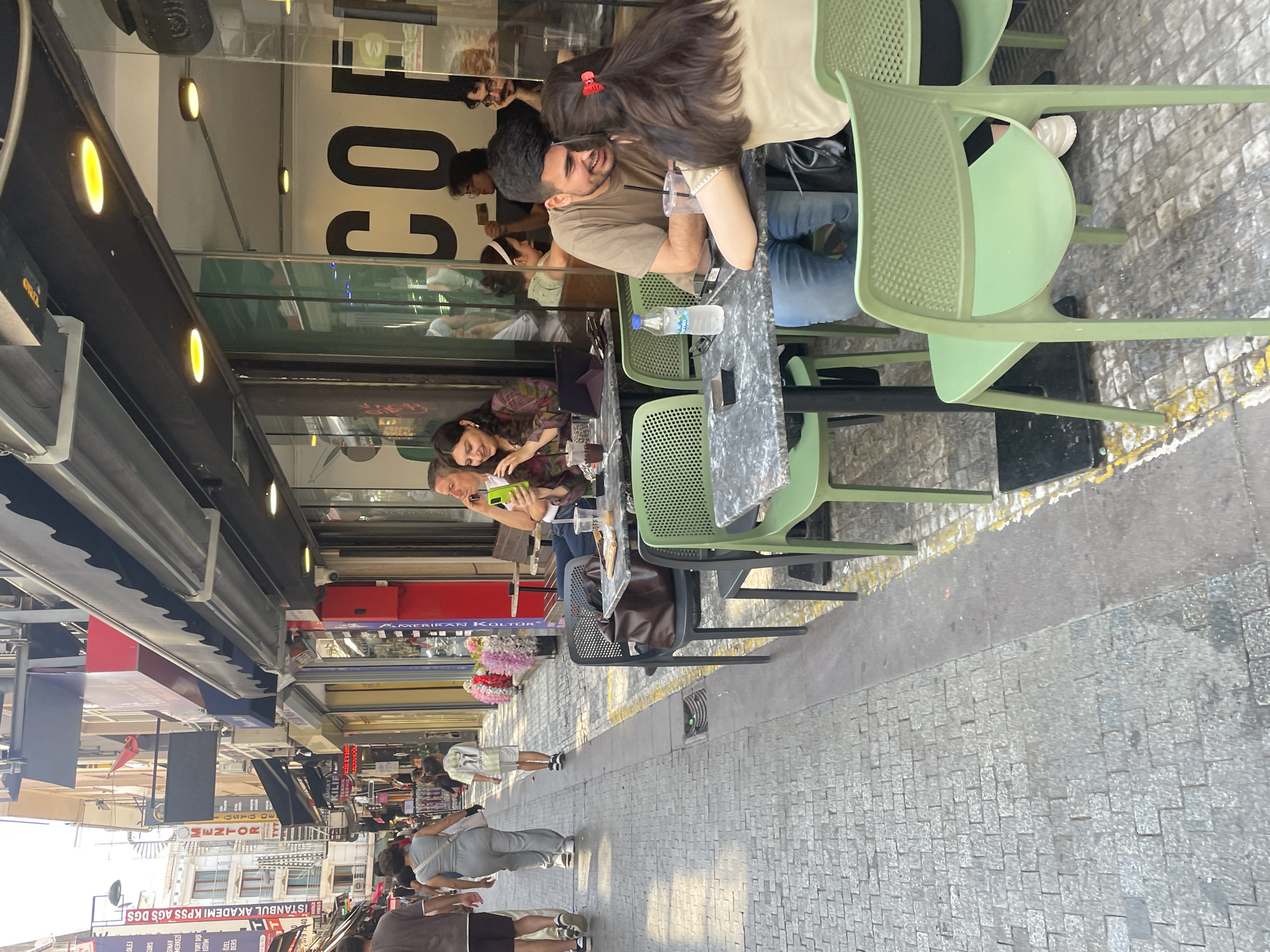 The image depicts a street scene in Kadıköy, Turkey, likely on Bahariye Caddesi, a pedestrian-friendly street known for its shops and cafes. A cobblestone sidewalk lines the left side of the frame, where several people are walking. In the foreground, a man and a woman are seated at a table outside a cafe. They appear to be in conversation, with smiles on their faces, and are leaning towards each other. A bottle of water and drinks are on the table, along with a phone.
The cafe has large glass windows that reveal the interior, where other patrons are visible. The cafe's exterior features awnings and signage, typical of businesses in this bustling urban area. The architecture suggests a European influence, common in Istanbul's older districts. The overall atmosphere is one of a relaxed, sunny day in a lively city.