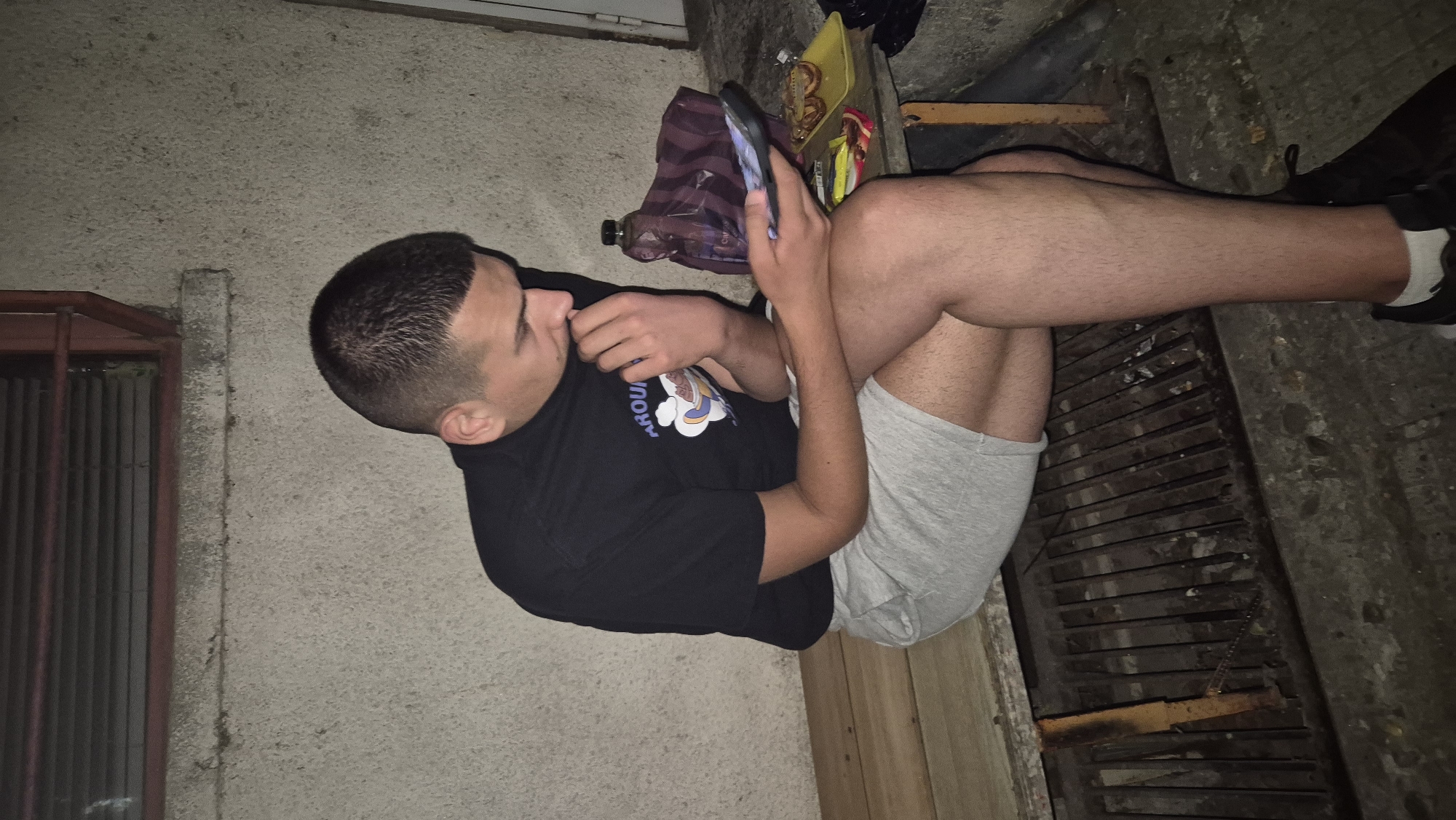 A young man sits on a wooden bench in an outdoor urban setting, possibly in Lovech, Bulgaria. He is wearing a black t-shirt with a graphic print and light grey shorts. He is holding a mobile phone in his right hand and appears to be looking at it, with his left hand near his mouth. Beside him on the bench, there are several items, including a purple bag, a plastic bottle, and a yellow tray with snacks. The bench is positioned over a metal grate. The background consists of a concrete wall and a section of blinds. The lighting suggests it is evening or night.