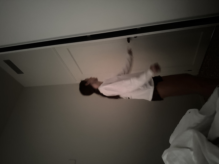 The image displays an interior room in Venice, Italy, characterized by subdued lighting. A young individual, possessing long dark hair tied in a ponytail, stands in motion slightly right of the frame's center. They are dressed in a light-colored, long-sleeved shirt and dark shorts. The person's head is turned to the right, directing their gaze toward a partially open white door occupying the right side of the frame. Their left arm is extended forward, indicating an interaction with the door. The walls are a neutral, light color. A rectangular ventilation grate is affixed near the ceiling in the upper right. The floor presents a speckled, patterned surface. In the lower-left foreground, crumpled white bedding is visible.