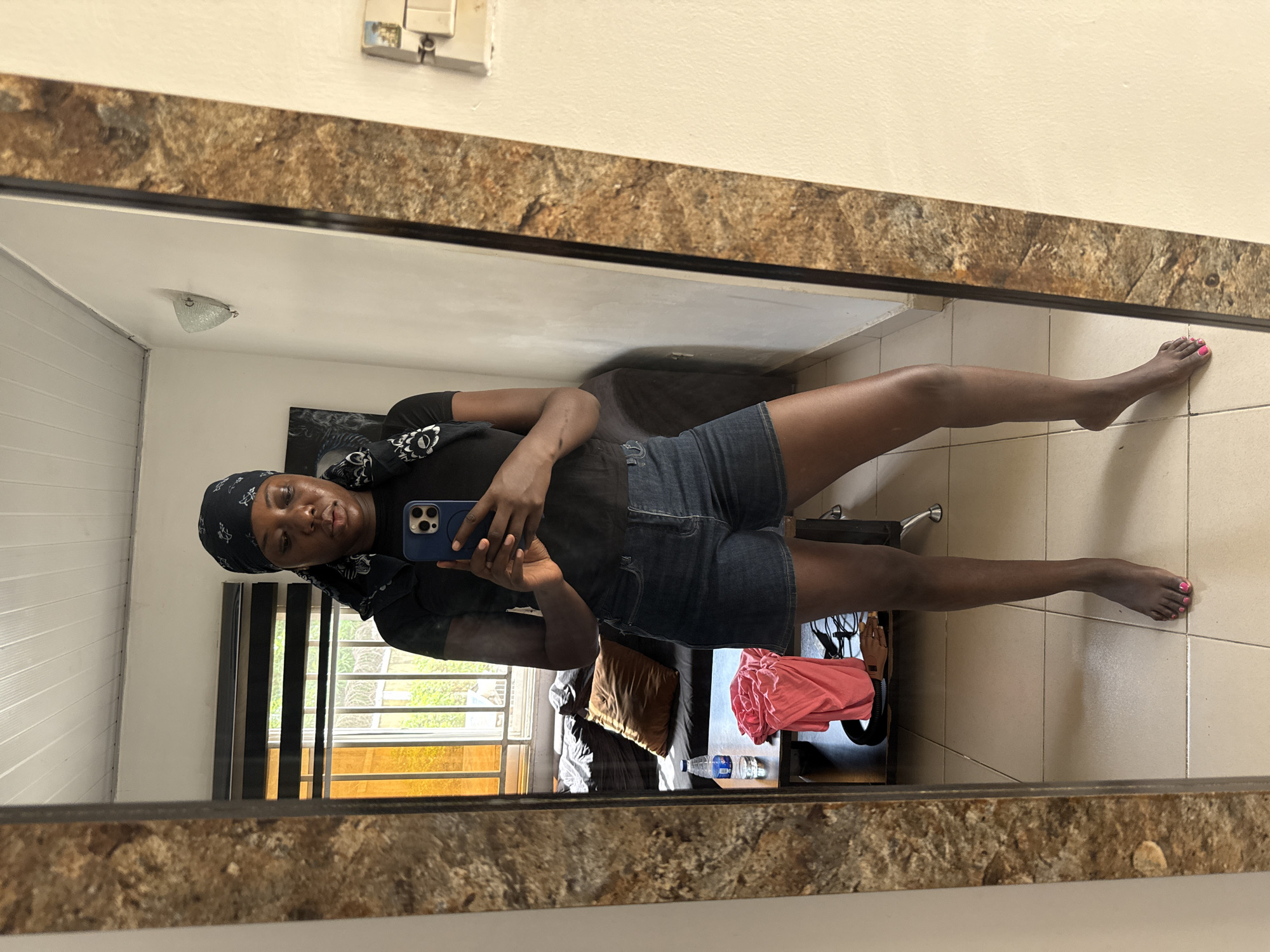 A person is reflected in a mirror. The person is Black, wears a headscarf, black t-shirt, and dark denim shorts, and holds a blue cell phone. The person has painted their toe nails pink.
The room reflected in the mirror contains a window with blinds, various items of clothing on a chair, and a floor tiled in light beige. The location is possibly in Abuja, Nigeria.