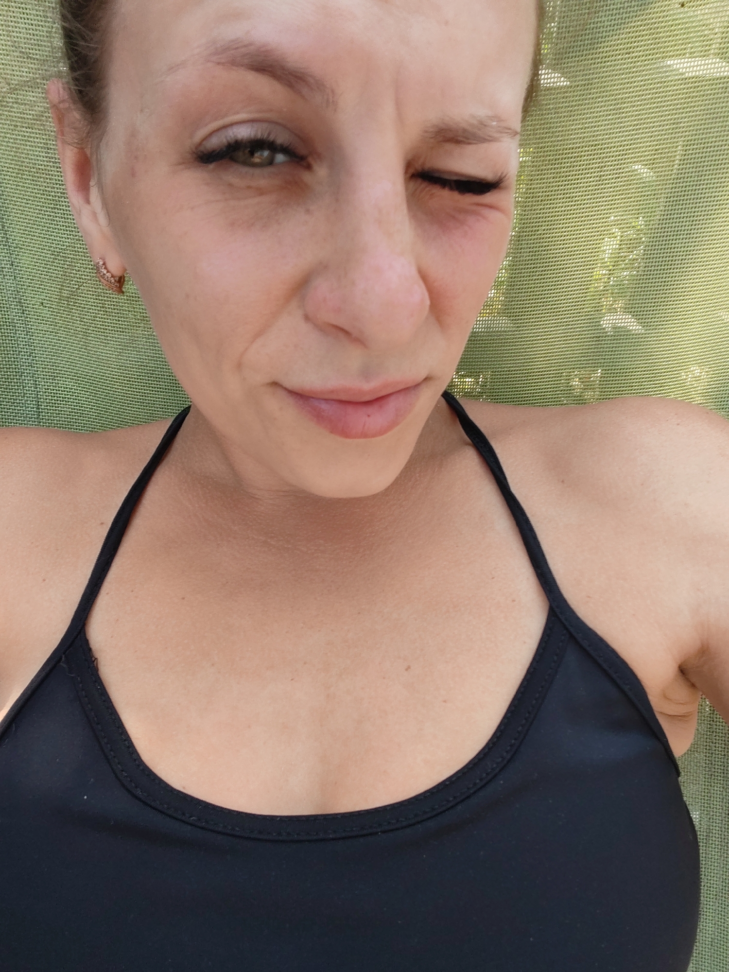 The image is a close-up, overhead shot of a woman lying down, with her upper body and face dominating the frame. She is wearing a black bikini top. Her right eye is open and looking towards the camera, while her left eye is closed in a wink. She has fair skin with some freckles, and her hair is pulled back. Her expression appears somewhat strained or pained, with a slight frown and a wrinkle between her eyebrows. The background is a textured green surface, possibly a lawn chair or umbrella, with some blurry foliage visible. There is no information in the image to suggest the location is Mezhdurechensk, Russia.