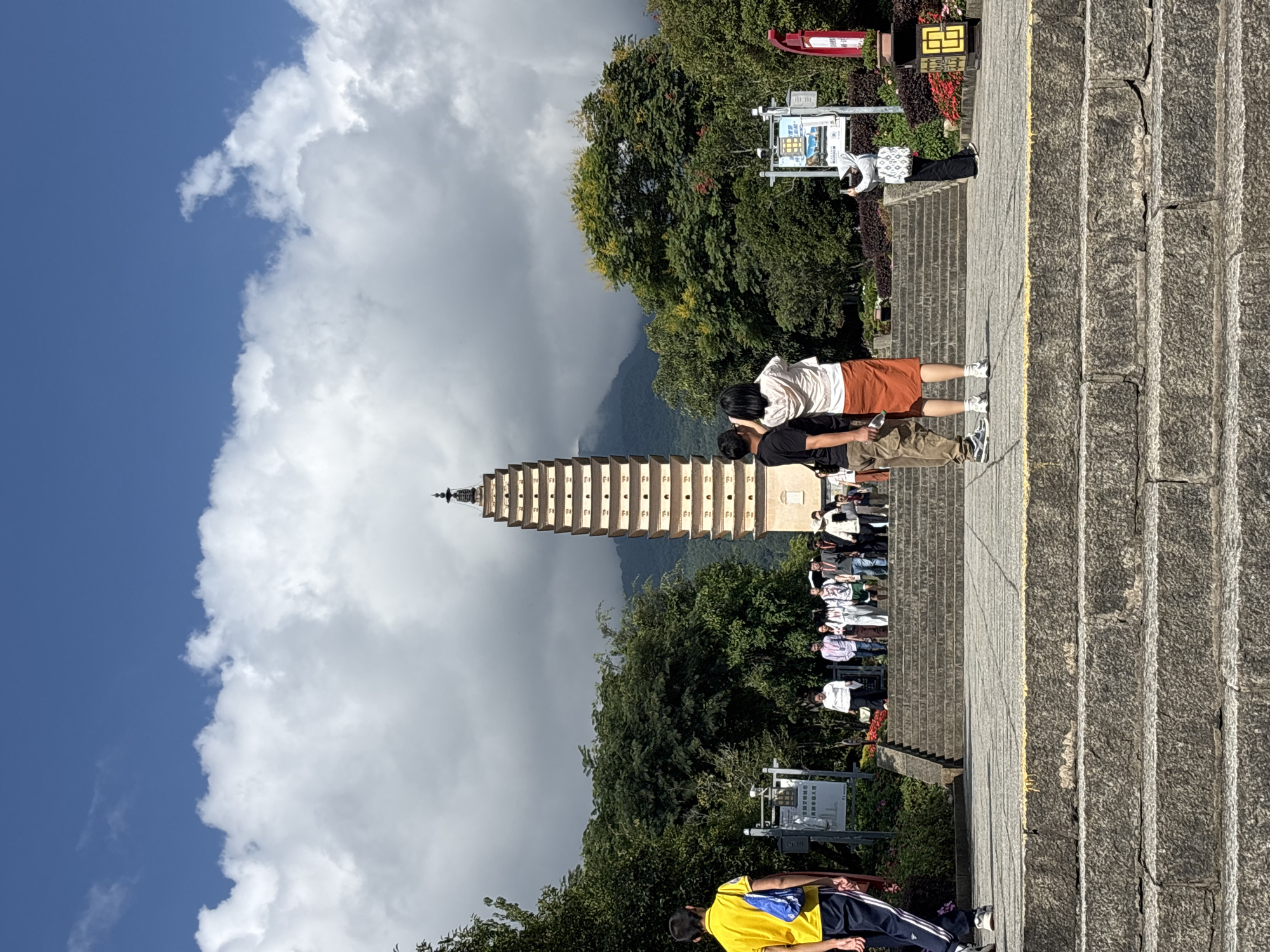 In Dali City, China, a tall, multi-tiered pagoda stands prominently in the background, its tiered structure reaching towards a sky filled with dramatic white clouds against a clear blue expanse. The pagoda is situated amidst lush green trees, with the faint outline of a mountain visible behind it. In the foreground, a stone staircase leads up to a paved area where several people are gathered. Two individuals, a man and a woman, stand facing away from the camera, looking towards the pagoda, with the man in a dark t-shirt and khaki pants and the woman in a white top and orange skirt. Another woman, wearing a white hijab and black pants, is taking a photo with her phone. A group of tourists are ascending the stairs on the left side of the image, while various signs and decorative plants are visible on either side of the path. A person in a yellow shirt walks away from the camera on the left. The overall scene depicts a popular tourist destination with a focus on cultural landmarks and natural scenery.