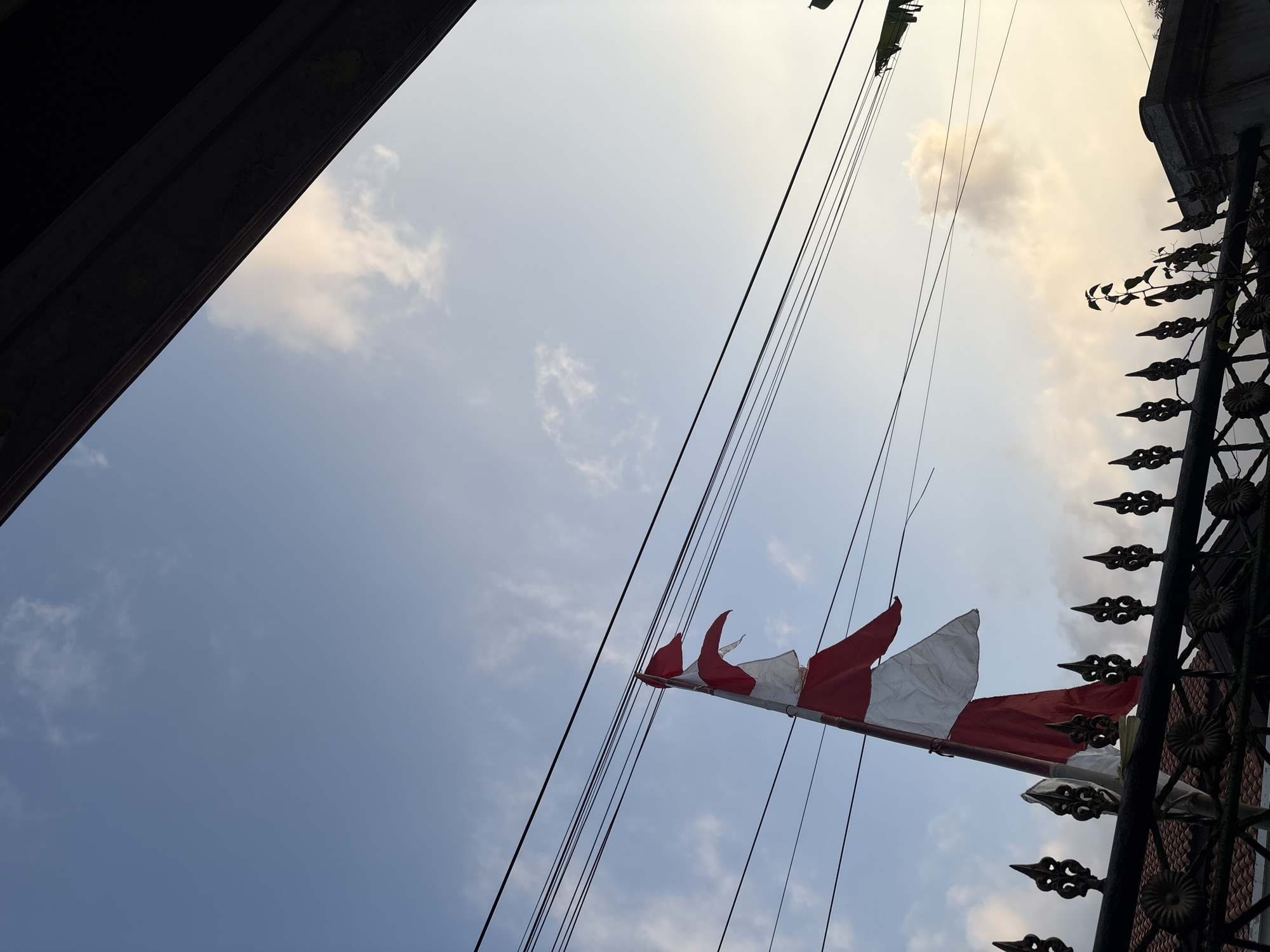 The image depicts a partially tattered Indonesian flag waving in the wind against a blue sky with scattered clouds. The flag is red and white, with the red stripe at the top and the white stripe at the bottom. It is attached to a pole and appears to be in an outdoor setting, possibly in front of a building. Several utility wires run horizontally across the frame, obscuring the sky in the upper portion of the image. A decorative metal fence with pointed finials is visible in the foreground, with a tiled roof and a structure behind it. The overall lighting suggests it might be late afternoon or early morning, given the soft glow in the sky. The scene is likely located in the city of Palimanan, Indonesia, given the presence of the Indonesian flag and typical urban elements like utility wires and fences.