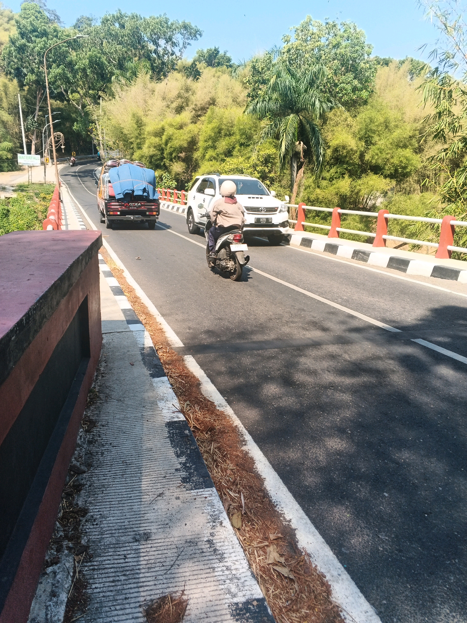 The image depicts a road scene on a sunny day, likely in a hilly or mountainous area, possibly outside the dense urban center of Semarang, Indonesia. The foreground shows a concrete walkway with a dark red and black striped wall on its left. The road itself is paved asphalt, marked with white lines. A motorcyclist wearing a helmet and a brown jacket is riding away from the camera, in the middle lane of the road. To the left of the motorcycle, a flatbed truck with a blue tarp covering its cargo is also moving forward. To the right of the motorcycle, a white SUV is traveling in the same direction. The road is bordered by a white and black striped barrier with red posts. The background is filled with lush green trees and bamboo vegetation, suggesting a natural or semi-rural environment. The sky is clear and blue. The presence of the road and vehicles indicates a developed area, but the dense foliage suggests it is not the heart of the city.