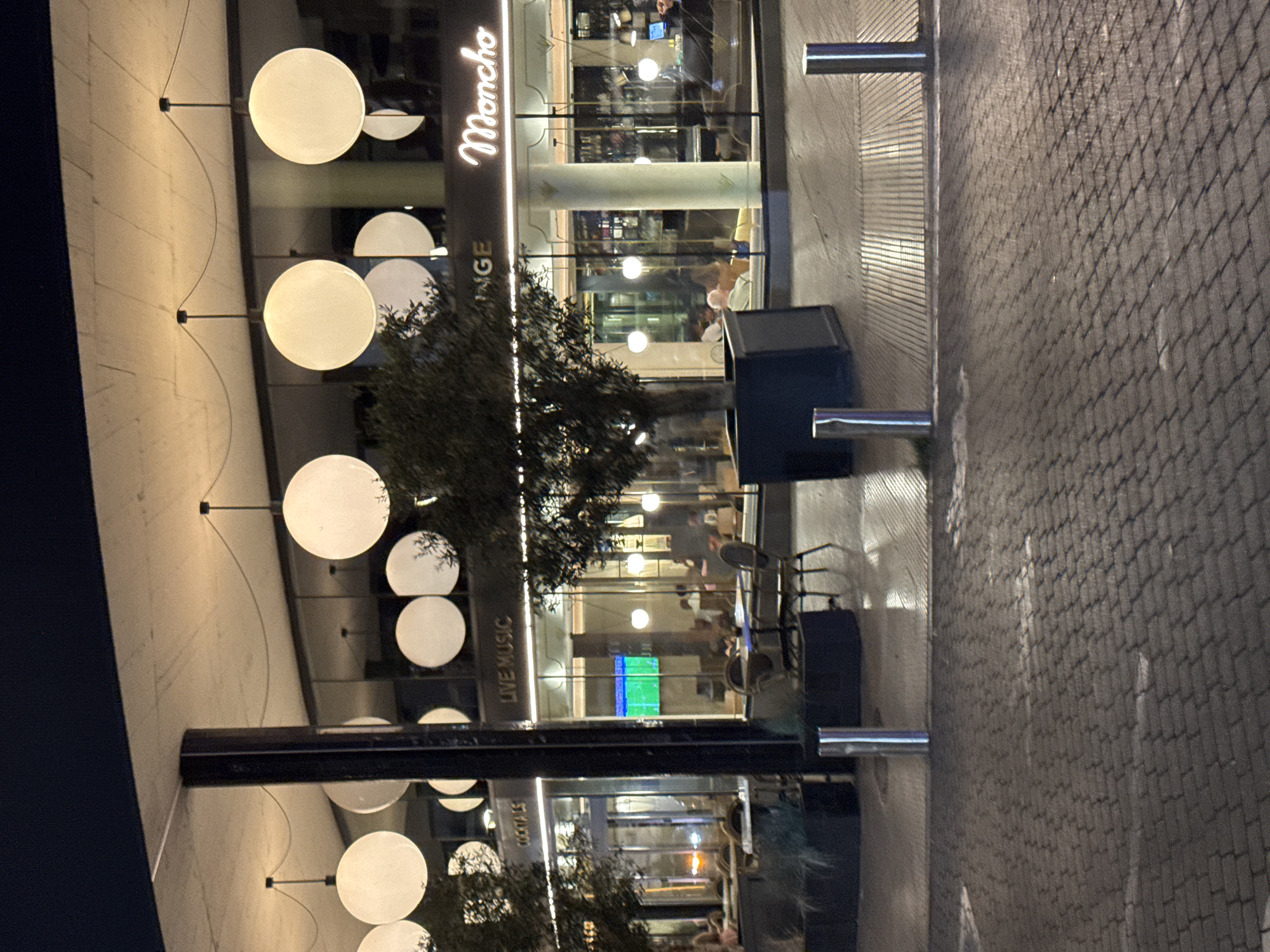The image depicts the exterior of a restaurant and lounge area at night, likely in a European city, with a prominent sign that reads "Moncho". The establishment features large glass windows, through which interior lighting and some patrons are visible. Inside, several tables and chairs are arranged, with some people seated. A large, modern overhead structure with spherical pendant lights is a dominant architectural feature. Trees are planted in planters outside the establishment, and the ground is paved with interlocking blocks. The scene suggests a lively, possibly upscale, dining and entertainment venue.