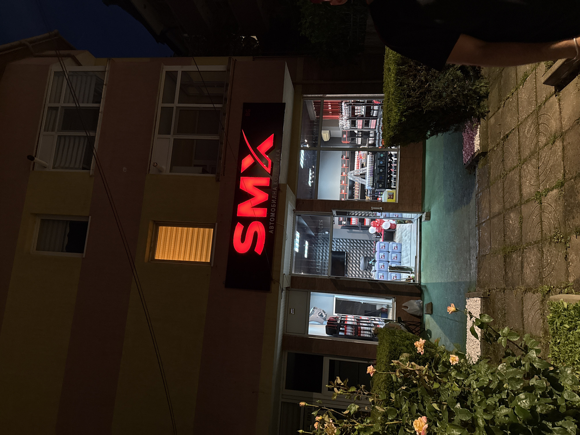 A two-story building in Targovishte, Bulgaria, is depicted at night, illuminated by artificial light. The building's facade features horizontal bands of light yellow and reddish-brown. Multiple windows are present on both floors; one upper-story window displays a warm orange glow emanating from behind closed blinds.
The ground floor hosts a commercial establishment, prominently identified by a red illuminated sign that reads "SMX" with a stylized 'X', and beneath it, "АВТОМОБИЛНА КОЗМЕТИКА" (Automobile Cosmetics). The shop's interior is visible through large glass windows and an open central doorway. Inside, extensive shelving units are stocked with various products, including items in red packaging and stacks of blue square containers on the floor. A white stepladder is visible. The interior also features a wall with a geometric pattern and a mounted flat-screen monitor. A cluster of red and white balloons is situated near the back of the shop's right side.
The exterior foreground includes a paved walkway composed of square concrete tiles, with weeds growing in the interstices. A patch of light green flooring or ground covering extends from the shop's entrance. To the left, a dense rose bush with several pale orange/peach roses in bloom occupies a garden bed, bordered by white decorative stones. A neatly trimmed green hedge lines the walkway on the right side. A small rectangular bed with pink/purple decorative stones is positioned in front of the hedge. Partially visible on the far right edge of the frame is the right shoulder and arm of a person, wearing a dark t-shirt and a silver bracelet on the wrist.