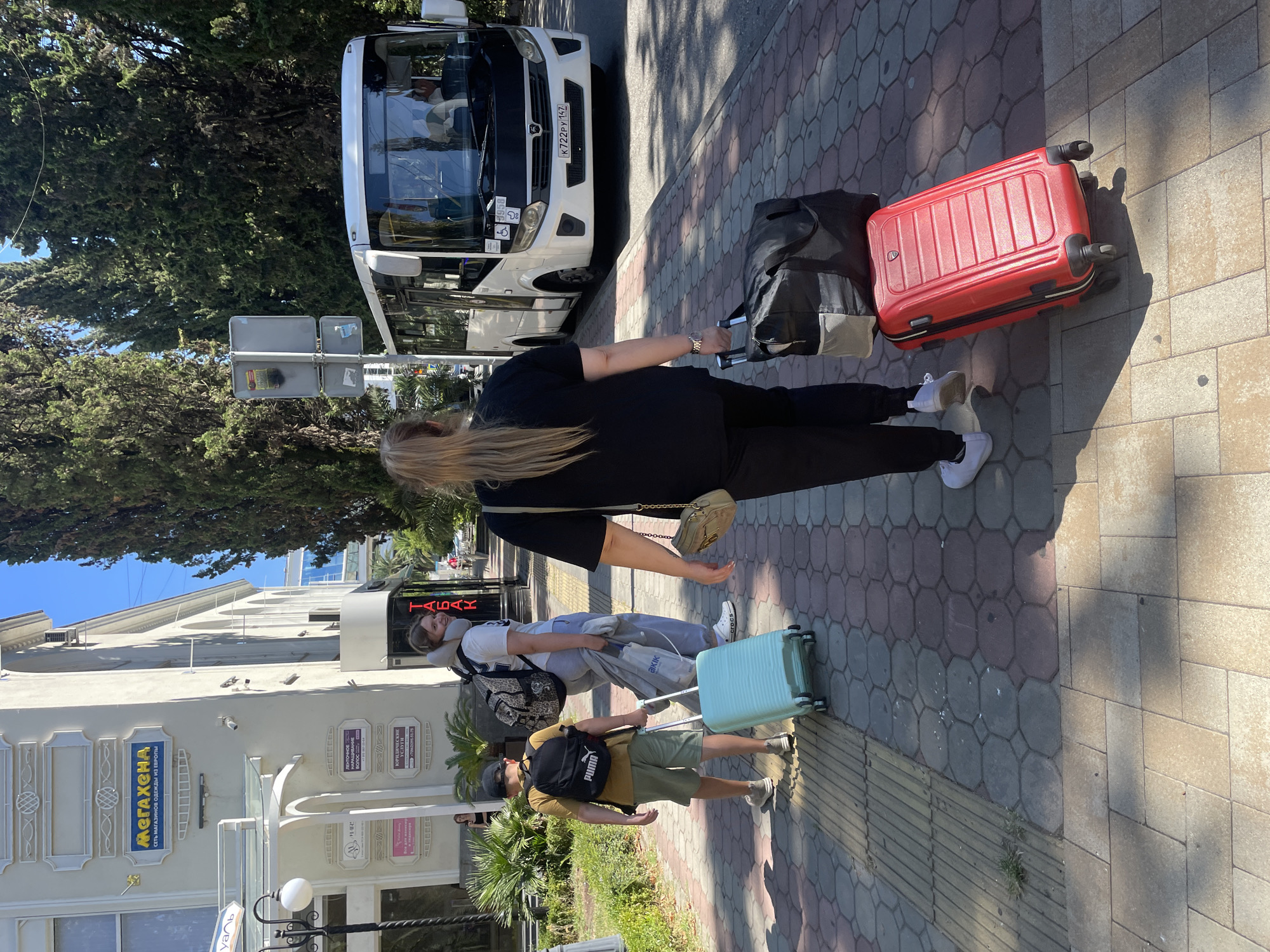 A group of tourists are walking on a paved sidewalk in Sochi, Russia, near a white bus. The scene takes place on a sunny day, with tall trees lining the street. A woman in the foreground, with her back to the camera, is pulling a red suitcase and a black bag on wheels. Behind her, a young woman with blonde hair is walking with a backpack and a light blue suitcase. A boy in a hat and backpack is walking in front of her, also pulling a light blue suitcase. The bus has a license plate that reads "K722PY 147". Buildings with signs are visible in the background.