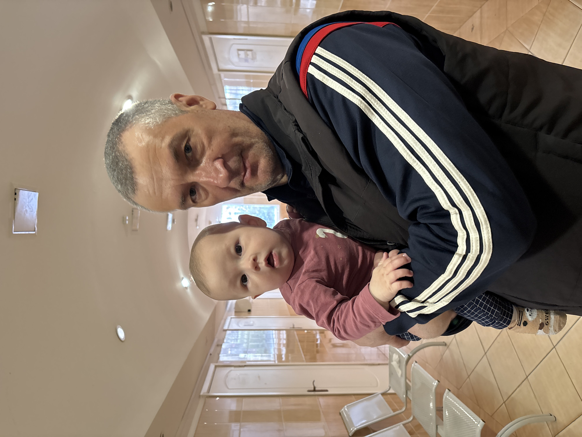 A man with graying hair is holding a baby in his arms. The man is wearing a navy blue tracksuit jacket with white stripes down the sleeves, red and blue accents on the shoulders, and a black vest over it. The baby is wearing a pink long-sleeved shirt and patterned leggings. The setting appears to be a waiting area with tile flooring and chairs. The image was taken in Sochi, Russia.