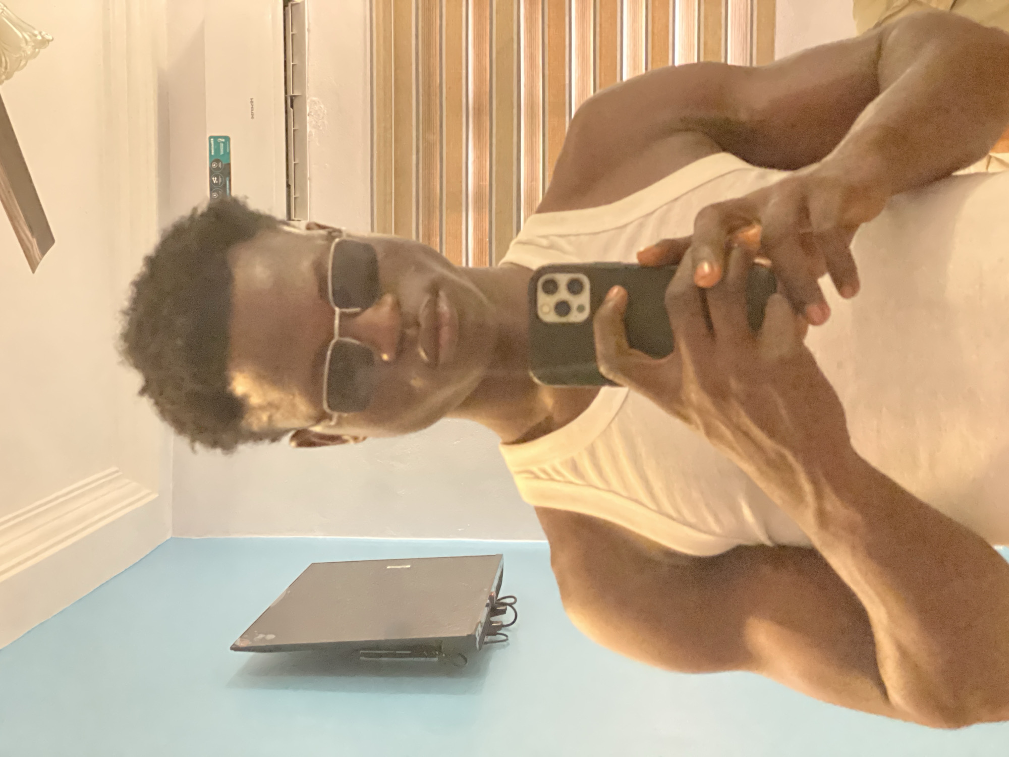 A dark-skinned man wearing sunglasses and a white tank top is taking a selfie with his smartphone. He is positioned in front of a mirror, reflecting the scene. A black laptop is placed on a blue floor near the wall. The background includes a white wall, a vertical air conditioner, and a slatted door. The location is suggested to be in Badagry, Nigeria.