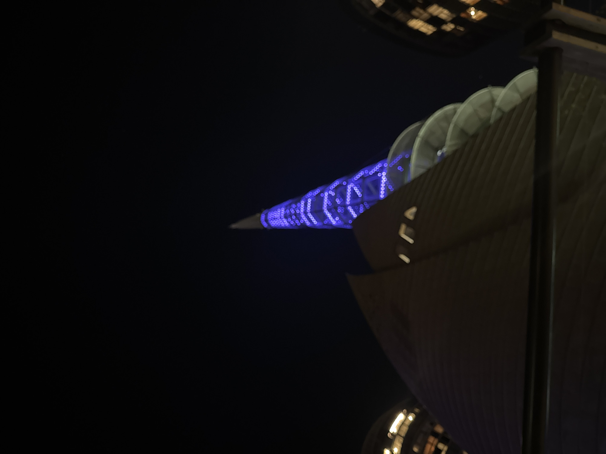 This night-time image from Perth, Australia, features a modern architectural scene dominated by a brightly illuminated spire and an adjacent building.
On the left, a long, slender, and conical structure extends diagonally across the frame, glowing with numerous distinct blue circular lights. These lights are intentionally out of focus, creating a bokeh effect, and appear to form intricate patterns, possibly abstract designs or stylized text, though specific characters are not clearly legible.
To the right, a large building or architectural facade rises, characterized by textured, possibly corrugated or ribbed, surfaces. Along its upper edge, a series of curved, shell-like or sail-like elements are visible, suggesting a unique design. This structure is dimly lit, appearing in muted tones against the dark background, contrasting with the vibrant blue of the spire. A dark, vertical support beam or pillar is positioned on the far right.
The sky is completely dark, indicating it is night. Blurry, warm-toned lights and reflections are visible in the upper and lower right corners, suggesting other illuminated parts of the environment or nearby structures. There are no people visible, and no specific activity is taking place other than the architectural illumination. The scene highlights the modern and distinctive design of the structures through their dramatic night lighting.