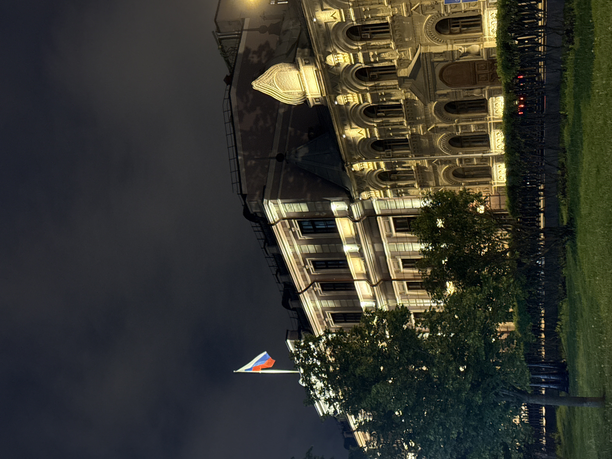 At night in Moscow, Russia, a building, likely the State Historical Museum, stands prominently against a dark sky. The Russian flag is visible on a pole, positioned in front of the building. A row of trees is situated in front of the building, adding a layer of greenery to the scene. A patch of grass stretches to the right of the trees. The building is illuminated, highlighting its architectural details.