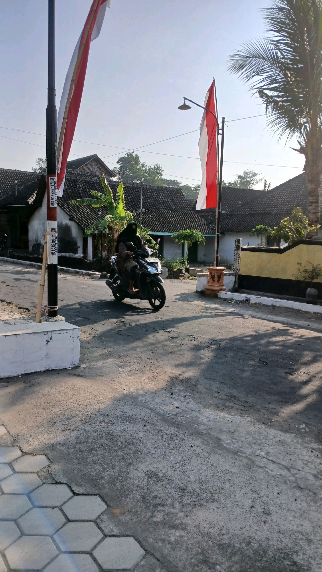 In the village of Geplak, Indonesia, on a clear, sunlit day, two individuals ride a black scooter along a paved road. The person in front, wearing a black hijab and dark clothing, is driving, while the passenger behind them, dressed in brown, is seated behind. The scooter is positioned in the middle of the road, with its front wheel turned slightly to the left. The background reveals traditional Indonesian houses with tiled roofs, some displaying a national flag of red and white. A palm tree stands to the right of the frame, its fronds casting shadows on the scene. To the left, a lamppost stands adorned with a red and white flag. The foreground shows a sidewalk made of hexagonal paving stones and a concrete barrier.