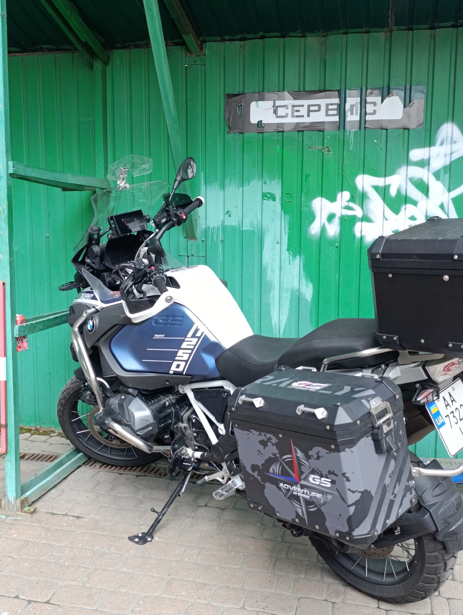 The image depicts a white and blue BMW R1250GS Adventure motorcycle parked on a paved surface in front of a green corrugated metal wall. The motorcycle is equipped with black panniers on both sides and a black top box. White and grey graffiti is visible on the green wall. A sign with the Cyrillic word "СЕРВІС" (service) is affixed to the wall. The motorcycle has a license plate with Ukrainian markings. The scene appears to be in an outdoor, urban environment, possibly a service area or parking lot in Kyiv, Ukraine. The motorcycle is stationary and no people are present in the frame.