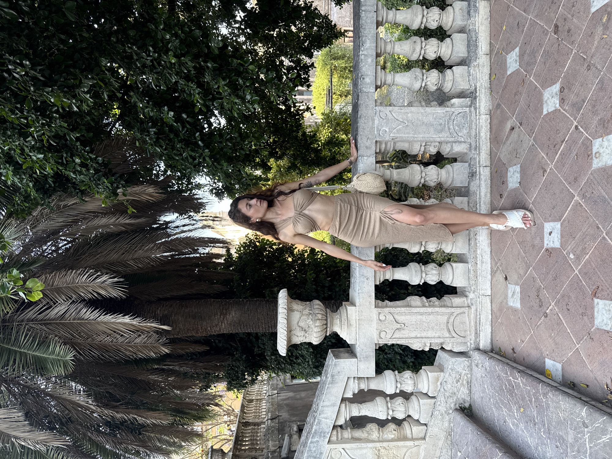 An outdoor, full-length portrait features a woman leaning against an ornate stone balustrade, likely within a historic garden or terrace in Catania, Italy. The woman is positioned in profile, facing left (viewer's right), with her right arm extended and resting on the balustrade, while her left hand is near her waist. She wears a light brown or tan two-piece outfit, including a crop top and a midi skirt with a high slit, paired with white open-toed sandals. A small, woven straw bag hangs from her right shoulder, and she has long, dark, wavy hair.
The setting is characterized by detailed stone architecture, including the balustrade with intricate carvings and stone steps below it. The ground is paved with a distinctive pattern of brown and white square tiles arranged diagonally. The background and foreground are filled with abundant green foliage from dense trees and prominent palm fronds, indicating a lush, warm environment. The scene is illuminated by natural daylight, suggesting it is late morning or afternoon on a clear day. No visible text is present in the image.