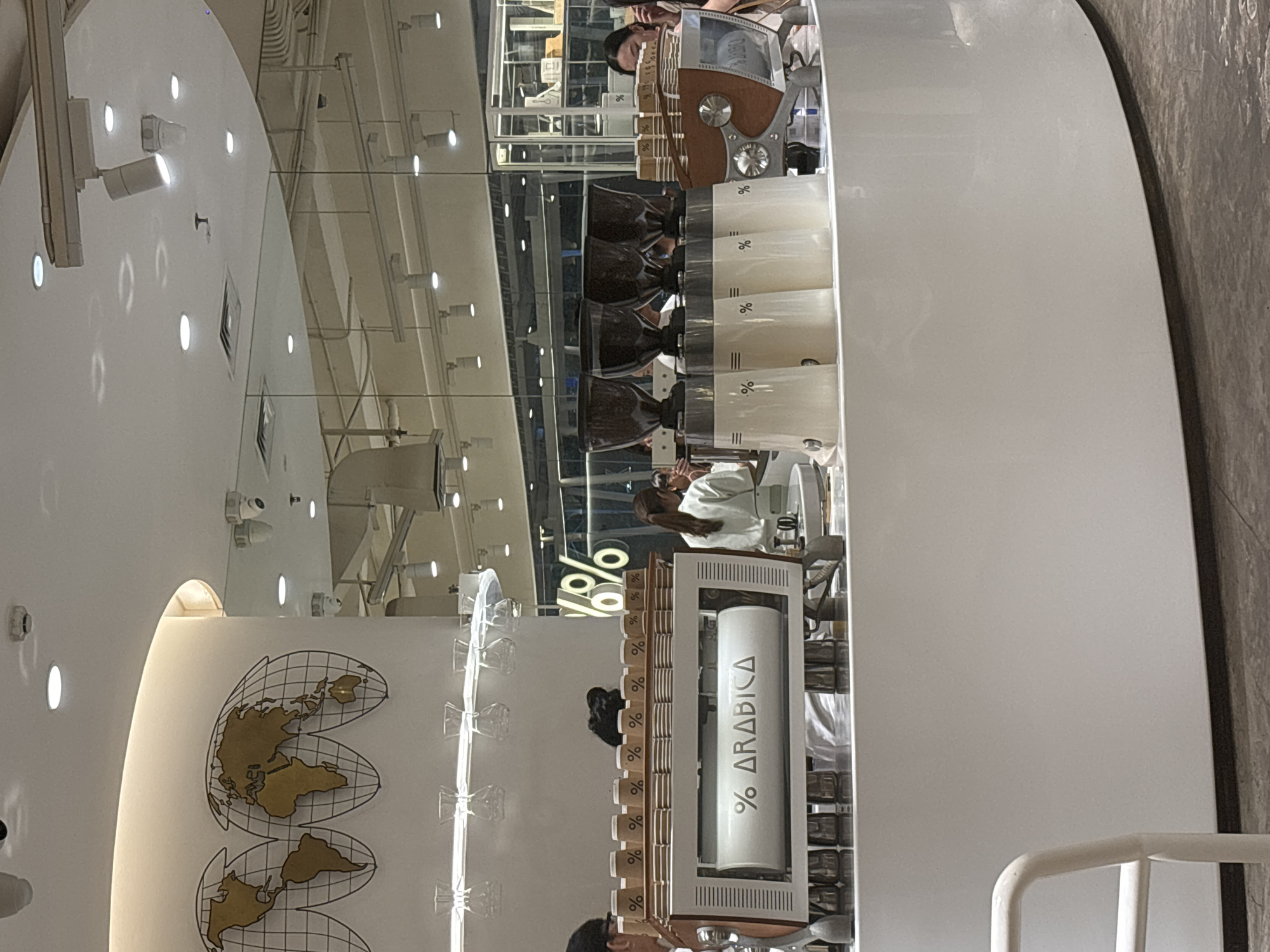 The image shows the interior of a % Arabica coffee shop in Kuta, Indonesia. A prominent % Arabica espresso machine with a white cylinder displaying the brand name and logo is in the foreground. Several coffee cups, also branded with the % logo, are lined up behind the machine. In the background, there are customers and staff members, with some individuals being served. The shop's interior features a clean, modern design with white walls and a polished floor. The ceiling has numerous light fixtures and a mirrored surface reflecting the shop's setup. Decorative world maps are mounted on a curved wall.