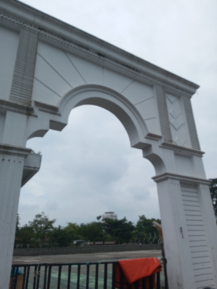 The image displays a large, white, classically-styled architectural archway located in Daringbadi, India, viewed from a low angle against an overcast sky. The archway features a semicircular opening. Its left supporting pillar is decorated with vertical fluting patterns, while the right pillar shows a series of chevron-like designs. Decorative molding runs along the top cornice and outlines the arch. Through the archway, a line of green trees is visible in the mid-ground, behind which a white building with a cylindrical or domed upper section stands in the distance. Below the trees, a dark, low wall or structure displays partially legible text. In the immediate foreground, a black metal railing with a dark green mesh infill is present. A folded orange object, possibly a life jacket, is draped over the right side of this railing. No individuals or specific actions are discernible.