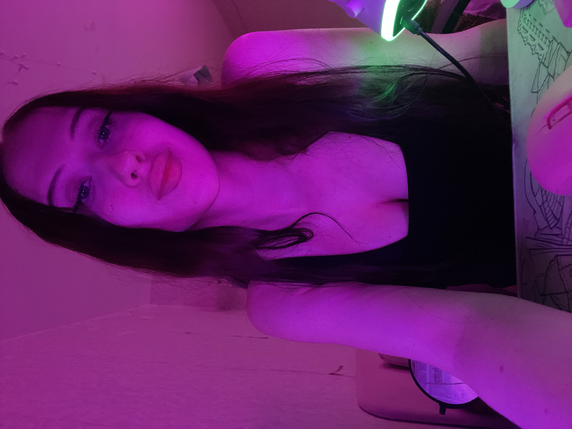 A young woman with long, dark hair and makeup, wearing a black tank top, is seated indoors in front of a computer setup. She is illuminated by pink and green lighting, casting purple hues on her skin and the surrounding environment. A microphone is visible to her left, and a computer mouse is in the foreground. The background appears to be a wall with some textured detail, and a portion of a chair is visible. The overall impression is that of a person engaged in online activities, possibly streaming or gaming, in a personalized, low-light setting. It is not possible to determine the specific location in Votkinsk, Russia, based solely on the visual information provided in the image.