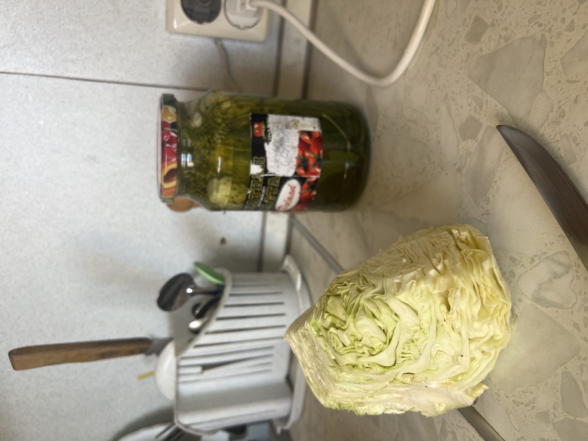An overhead shot shows a section of a quartered cabbage and a jar of pickles on a tiled kitchen counter. The cabbage, pale green and tightly packed, sits in the foreground. Behind it, a glass jar filled with pickles and brine is positioned to the right. A metal knife with a wooden handle lies on the counter in front of the cabbage, its blade pointing towards the viewer. In the background, a white utensil holder filled with cutlery and a knife is visible. A power outlet with a white cord plugged into it is mounted on the tiled wall behind the jar. The overall scene is lit by an overhead light, casting subtle shadows on the counter and wall.