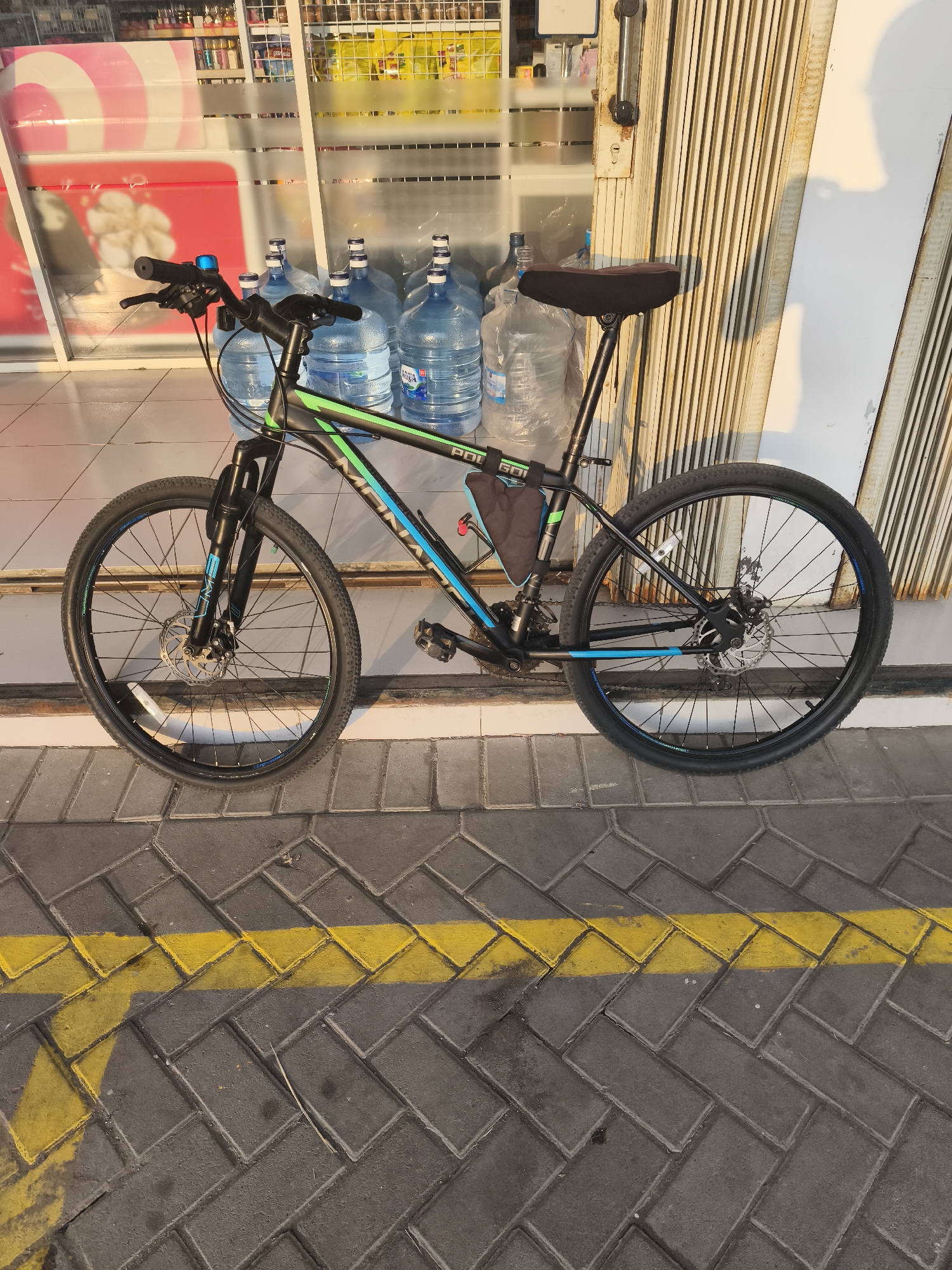 In the city of Kemiri Kidul, Indonesia, a black and green mountain bike is parked on a sidewalk. The bike is positioned horizontally, facing towards the right of the frame, with its front wheel to the left and rear wheel to the right. The bike has a black frame with green and blue accents. It is equipped with disc brakes on both wheels, a black saddle, and a small black bag attached to the frame. Behind the bike, a row of large water gallon jugs is visible, stacked near a storefront with metal rolling doors. The sidewalk is paved with grey interlocking bricks and marked with a yellow line. The lighting suggests it is daytime, with shadows cast by the bike and the surrounding structures.