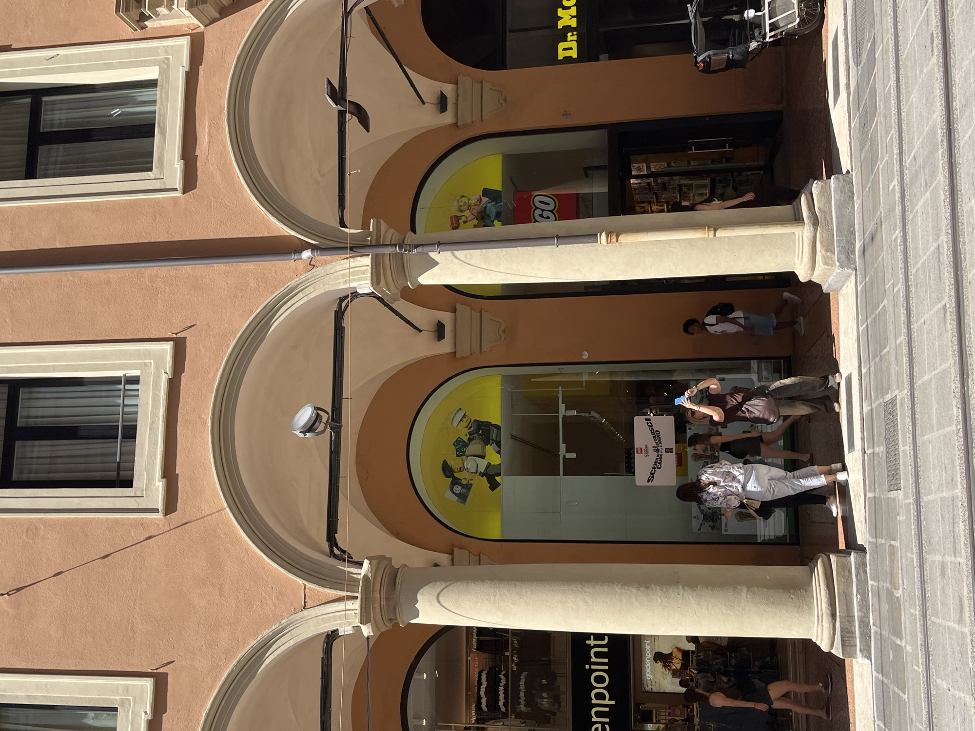 The image shows a street scene in Bologna, Italy, featuring a series of arched storefronts. The buildings are peach-colored with white trim around the windows. There is a LEGO store visible, indicated by the LEGO logo and displays in the windows. People are walking in front of the stores, some looking into the windows. A "goldenpoint" store is also visible. The architecture includes arched openings supported by pillars.