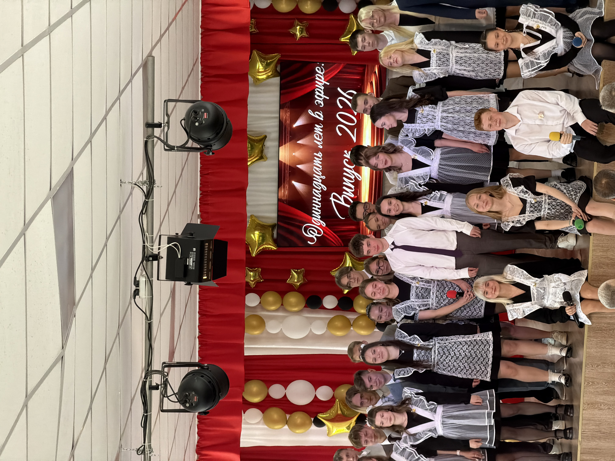 A group of young people, likely high school graduates, are standing together for a photograph in what appears to be an auditorium or event space in Izhevsk, Russia. They are positioned in front of a stage decorated with red and white curtains, gold and white balloons, and gold star-shaped decorations. There is a backdrop with the inscription "Одиннадцать лет в эфире. Выпуск 2026" which translates to "Eleven years on the air. Graduation 2026".
Most of the girls are wearing traditional school uniforms consisting of black dresses with white lace aprons and tights. Some are holding microphones. There is at least one boy wearing a white shirt and a tie.
Stage lighting equipment is visible on the left side of the image, including spotlights and a control panel. The overall environment suggests a celebratory event marking the end of high school.