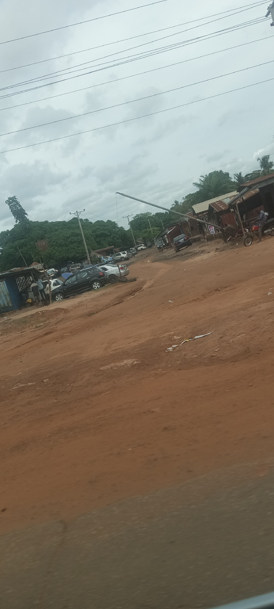The image depicts an unpaved road intersection in Markurdi, Nigeria. The road is primarily red dirt. Several vehicles are parked on the left side of the road. A group of people stand near a blue corrugated metal structure. In the background, a large tree is visible, along with a crane-like structure extending into the sky. On the right side, there are makeshift stalls with rusty metal roofs. A person sits on a motorcycle near one of the stalls. Electrical wires are strung across the sky. The sky is overcast and gray.
