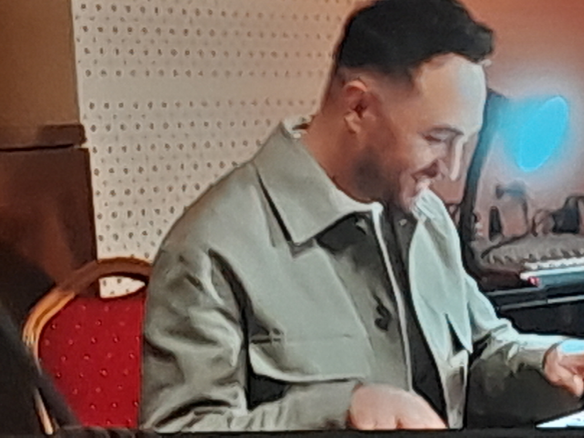 An adult male is seated in a chair, his body angled to the right, and he is looking down and to his right. He is wearing a light-colored collared jacket over a darker garment. The background consists of a textured wall and a red chair with a patterned seat. To the right of the male, a keyboard and a blue light source are visible. The man's right hand is resting on his leg, and his left hand is not visible.